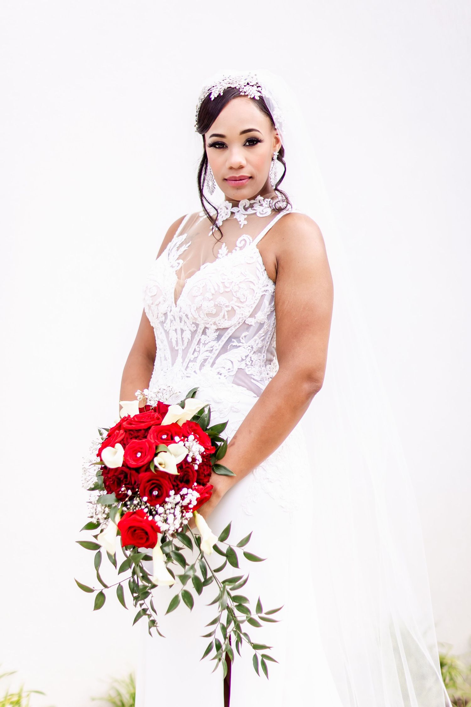 A bride in a detailed lace wedding gown posing with a red and white bouquet.