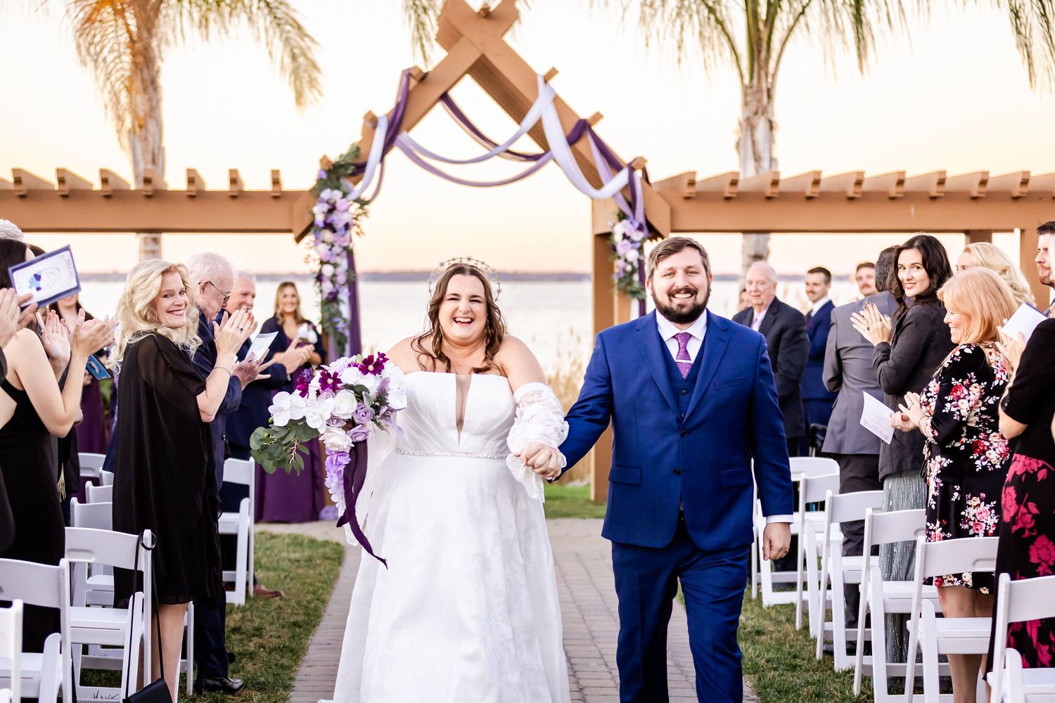 Wedding couple exits ceremony under arch with palm trees and cheering guests in background.