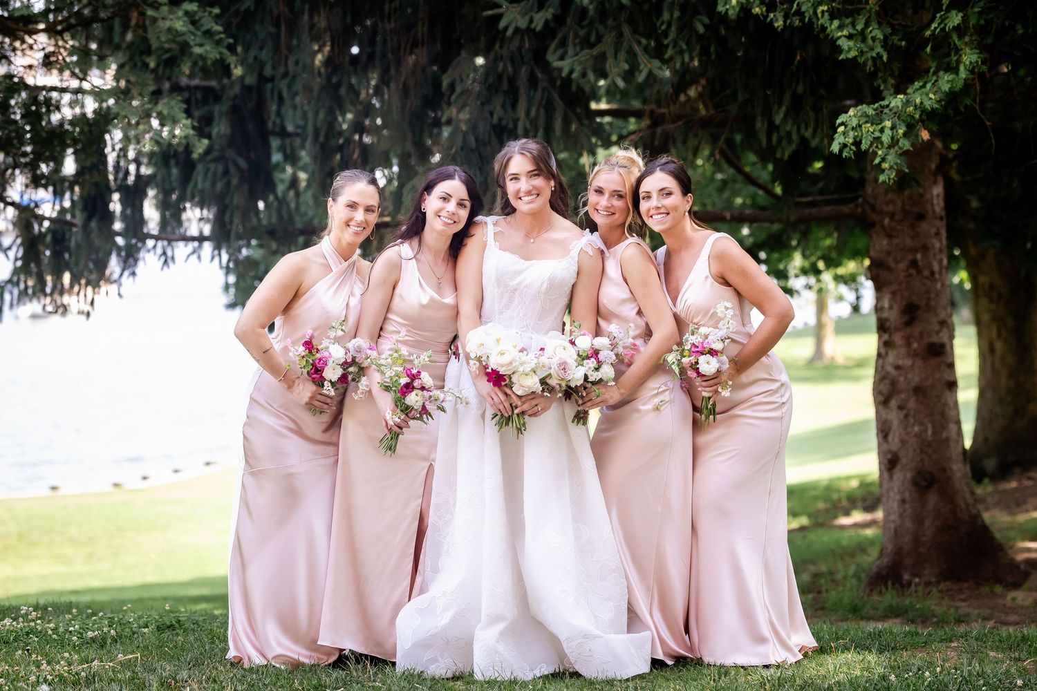 Bridesmaids in blush pink dresses pose together outdoors under trees.