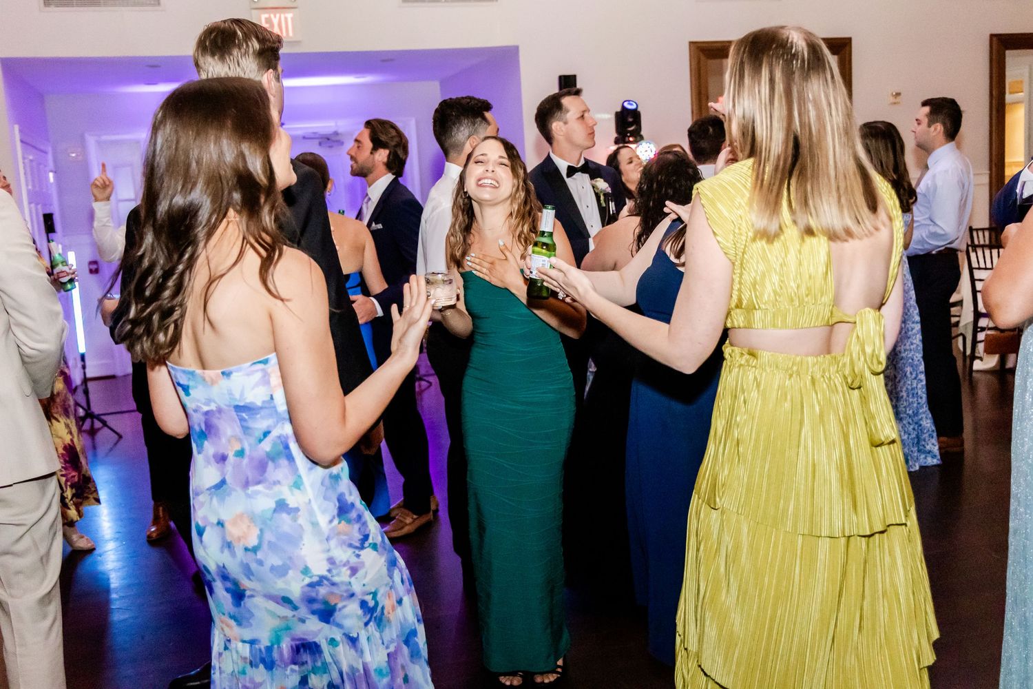 Wedding guests in colorful dresses dance together at an evening reception with purple lighting.