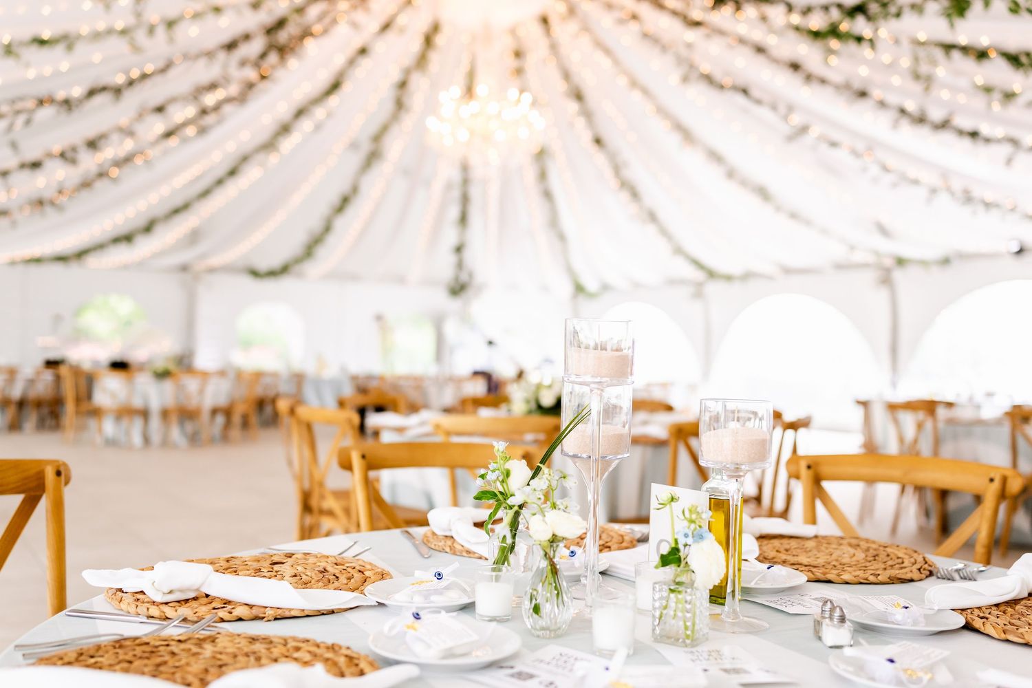 Wedding reception table settings with white linens and wooden chairs under draped fabric ceiling.