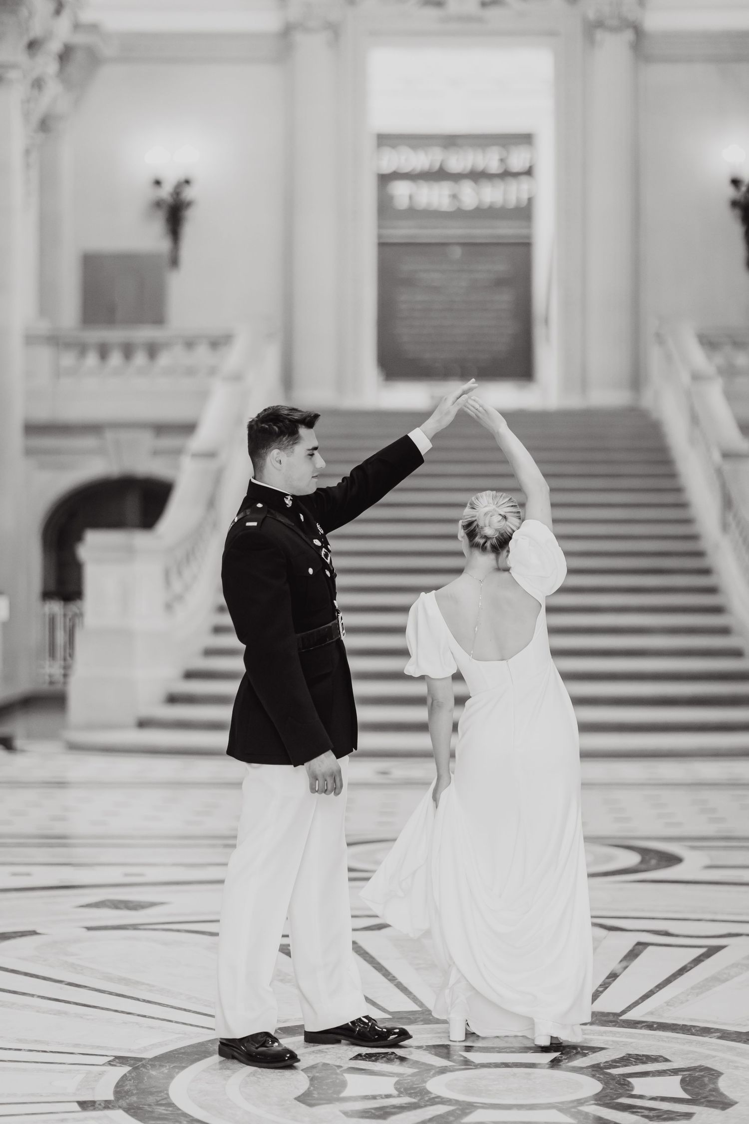 A couple in wedding attire dances together on an ornate marble floor in front of a grand staircase in black and white.