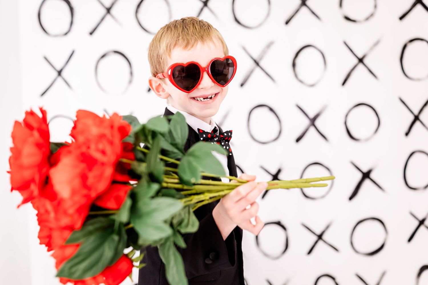Stylish dresser in heart sunglasses holds red roses against X's and O's backdrop.
