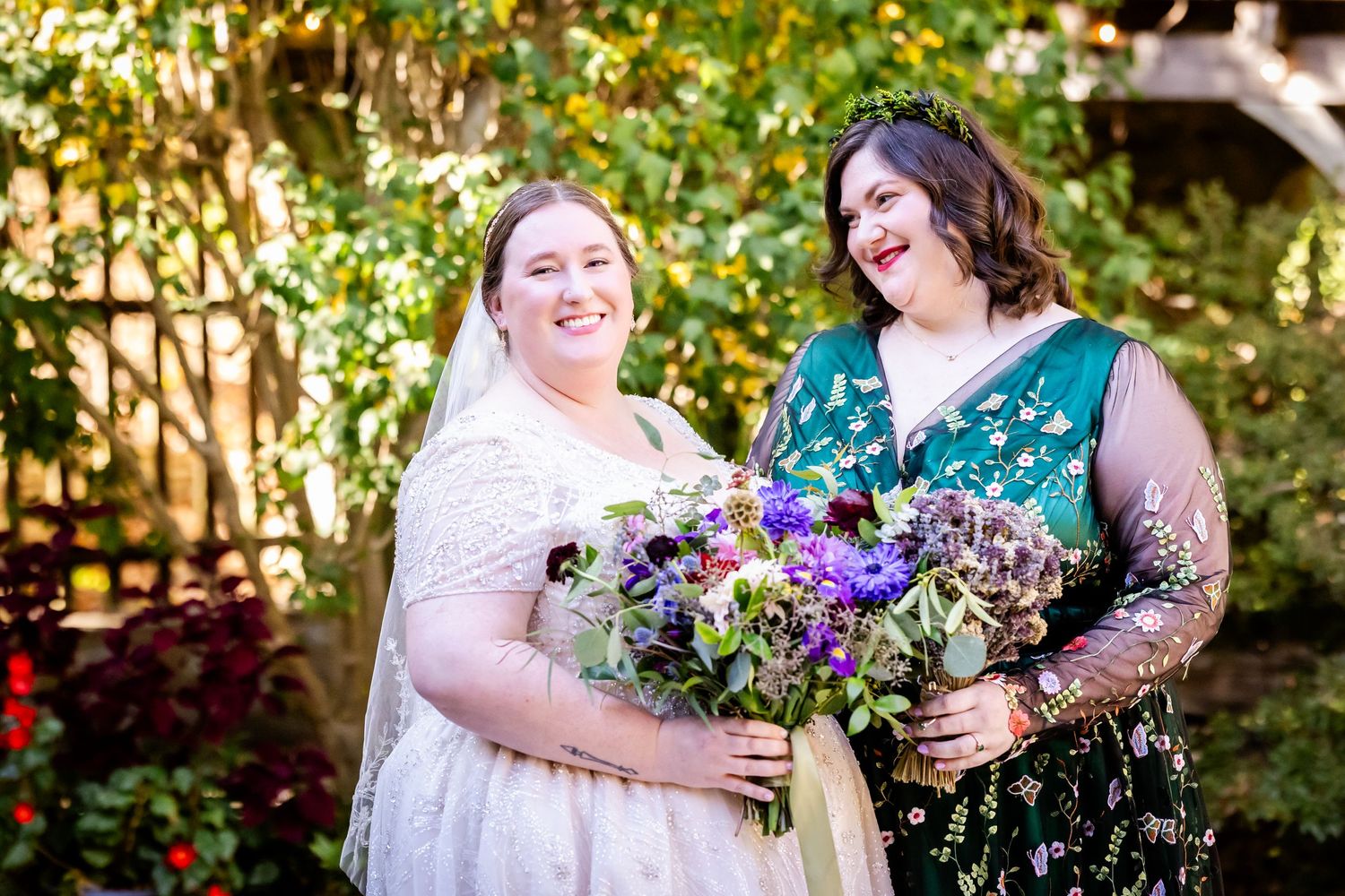 Two wedding party members share laughter while holding vibrant purple floral bouquets outdoors.