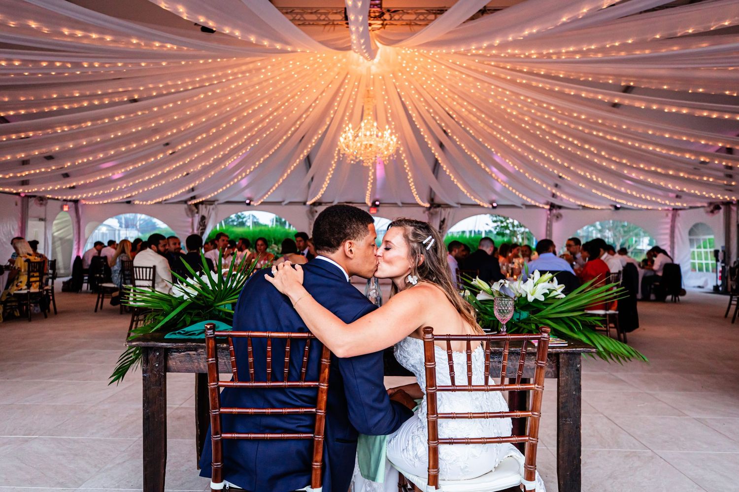 A couple shares a romantic moment under a stunning reception tent adorned with glittering string lights and chandeliers.
