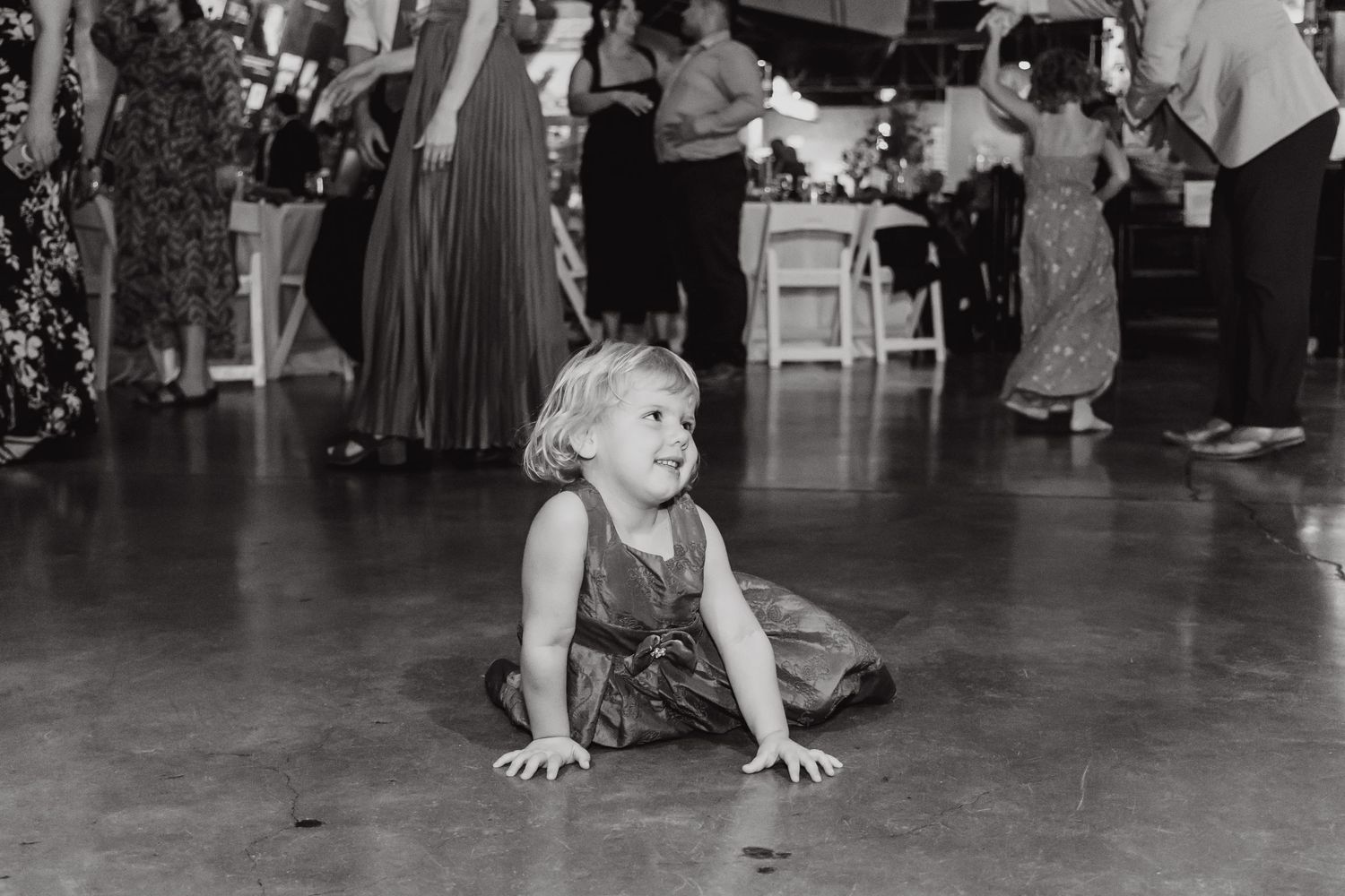 A small child in casual clothing sits on a dance floor during a celebratory event.