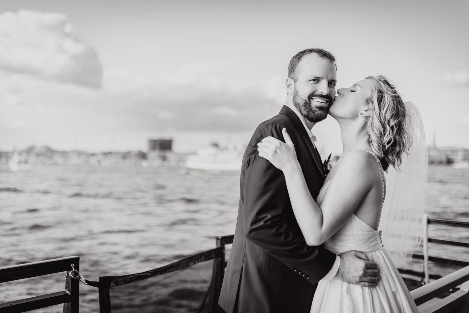 A couple embraces in formal wedding attire on a dock by the water in black and white.