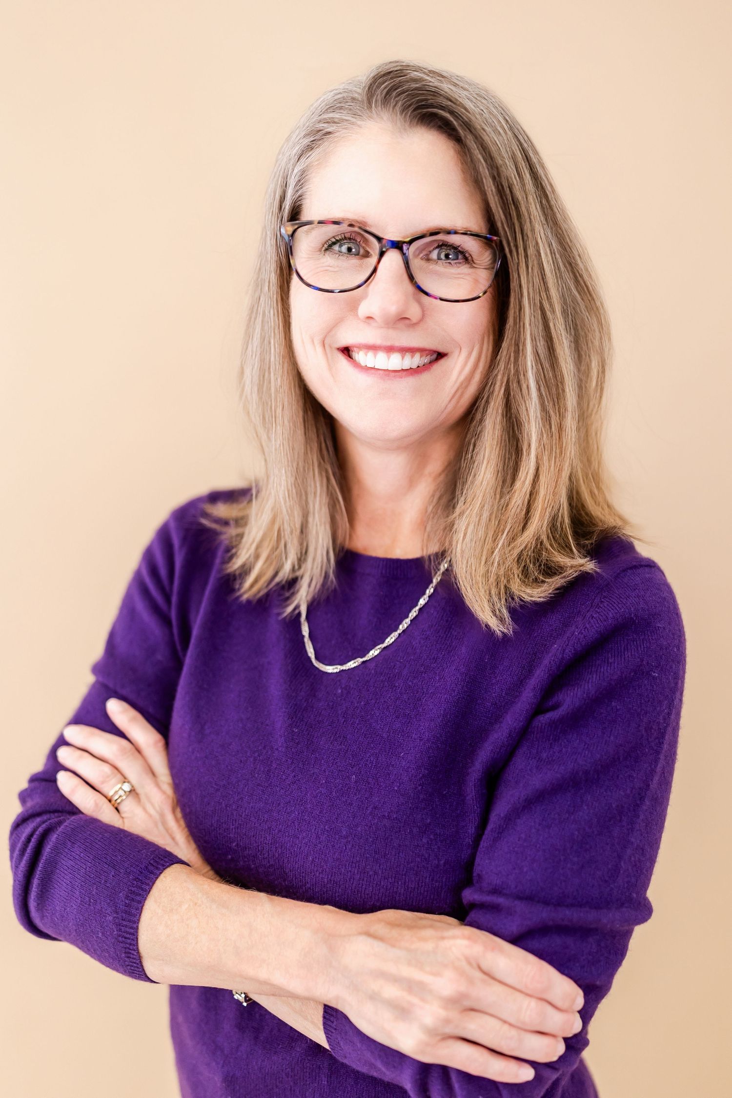 Smiling middle-aged woman with glasses and gray hair wearing a purple sweater, arms crossed, against a beige background.
