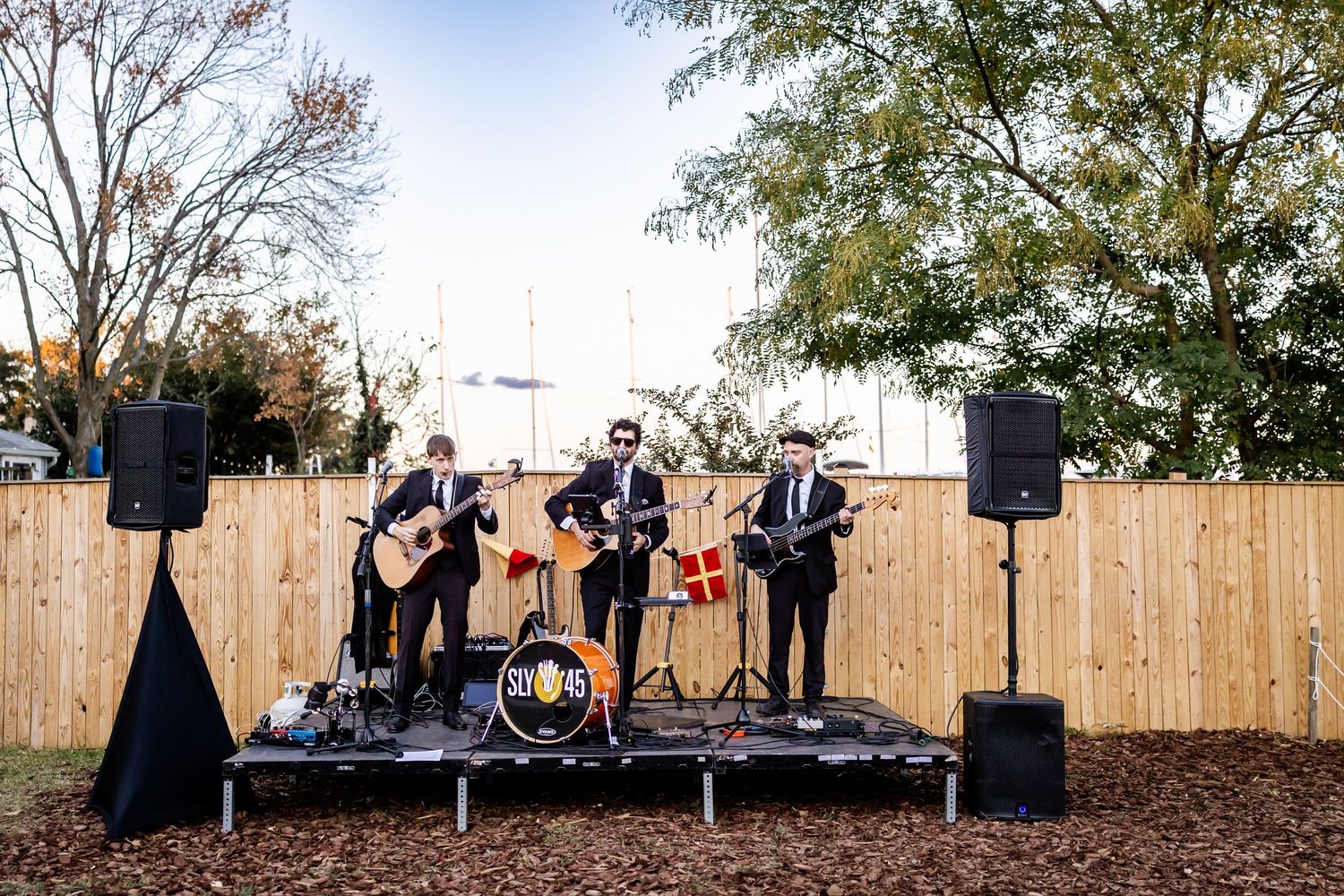 Four-piece band performing outdoors on a small stage with speakers, guitars, and drums against a wooden fence backdrop.