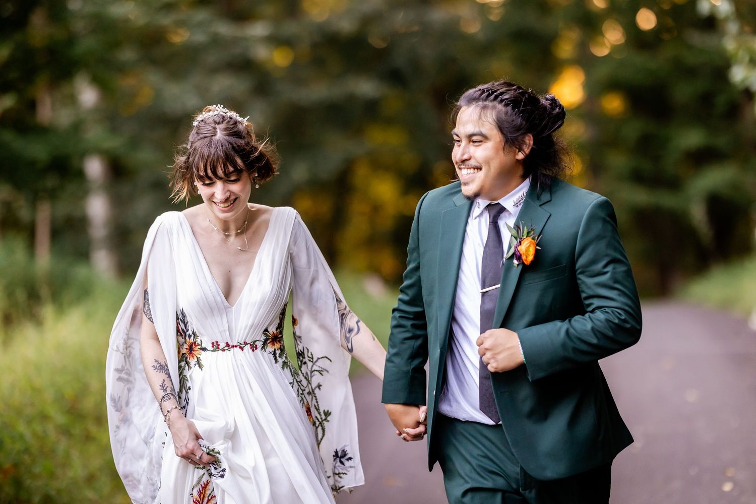 Newlywed couple laughing and walking together on a tree-lined path during an autumn wedding celebration.