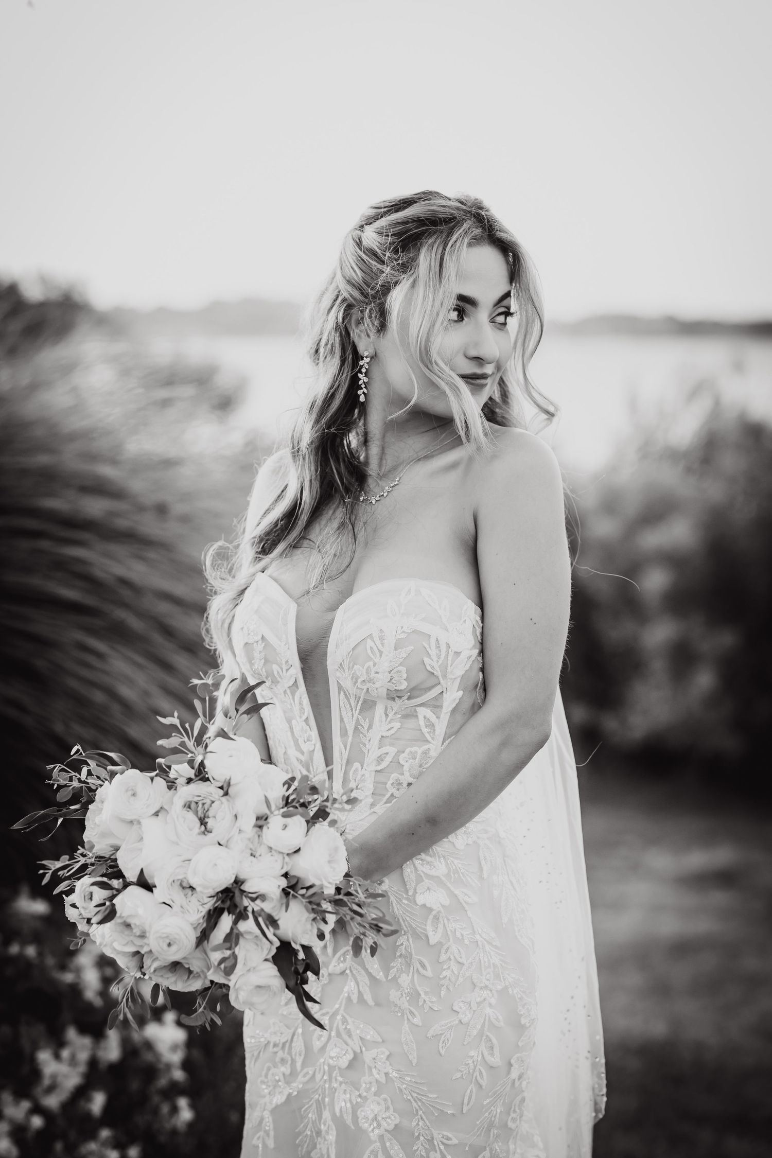 A bride in a white lace gown holds a romantic bouquet of flowers during sunset in black and white photography.
