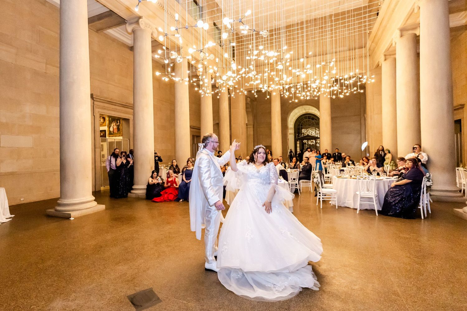 Newlyweds dance in an ornate ballroom with hanging crystal lights and grand columns.