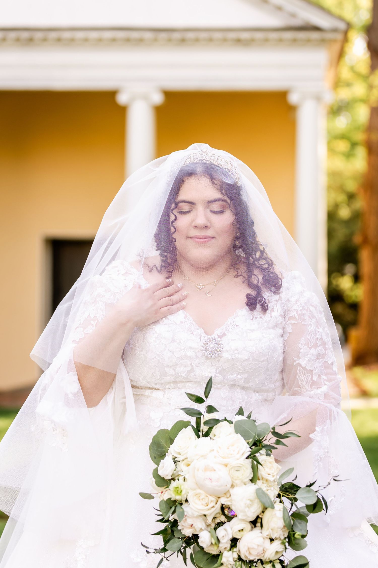 Bride in white lace gown holds elegant bouquet of white roses and greenery.