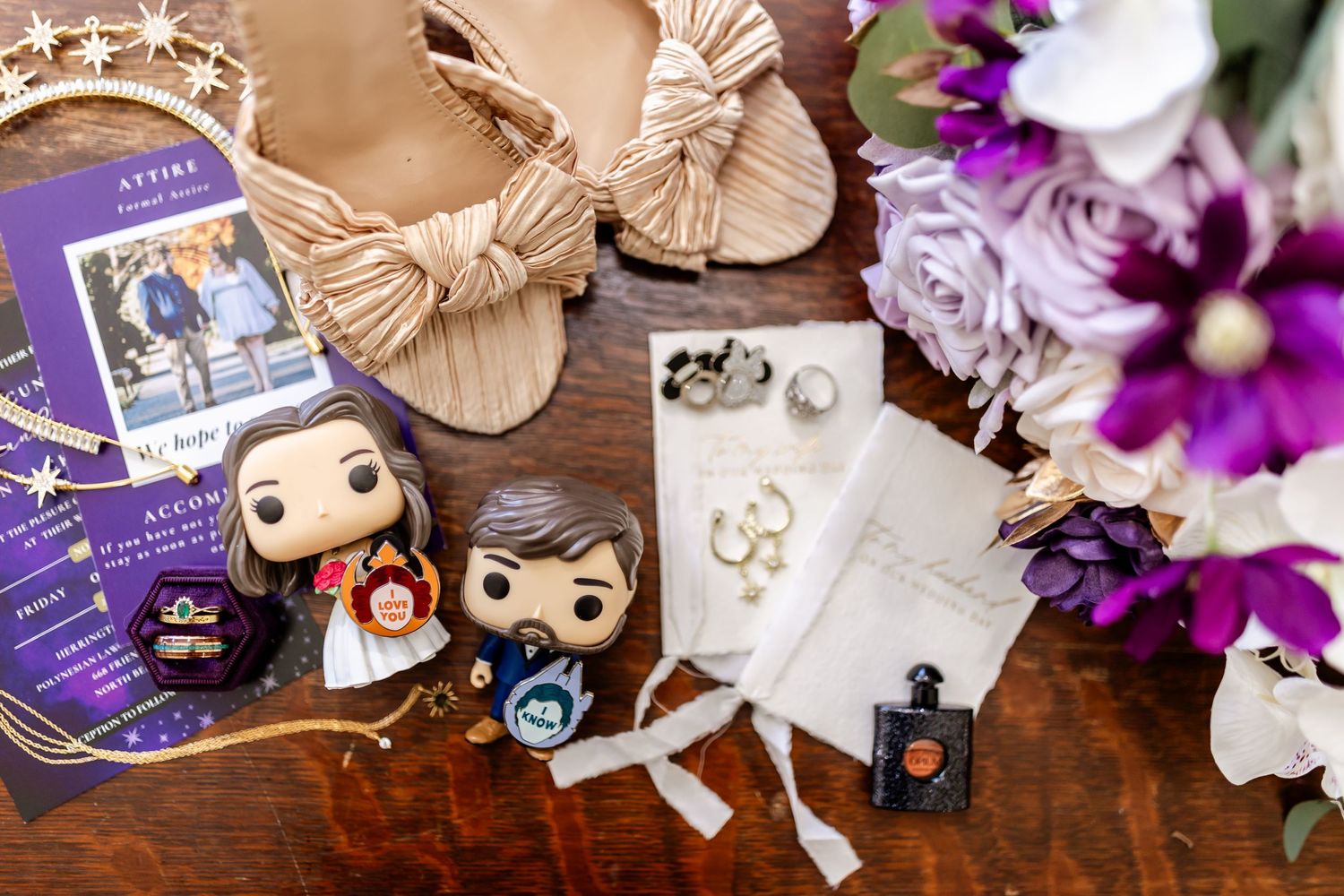 Wedding details flatlay with Funko Pops, invitations and purple flowers on wooden surface.