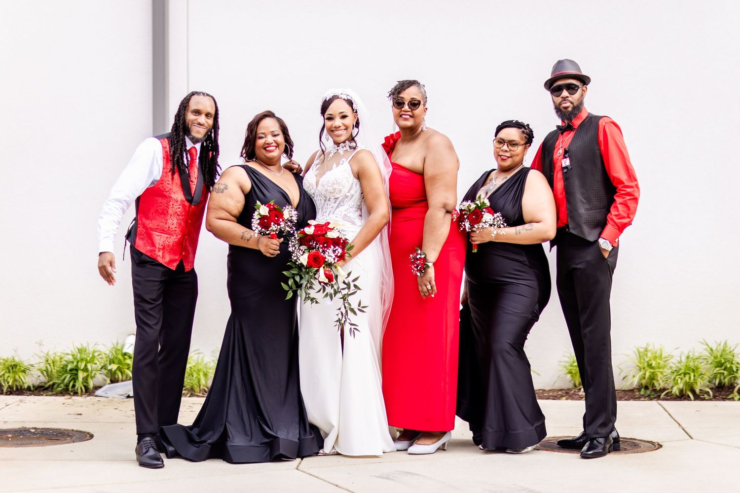 Wedding party in red and black formal attire pose together with bouquet against white wall.