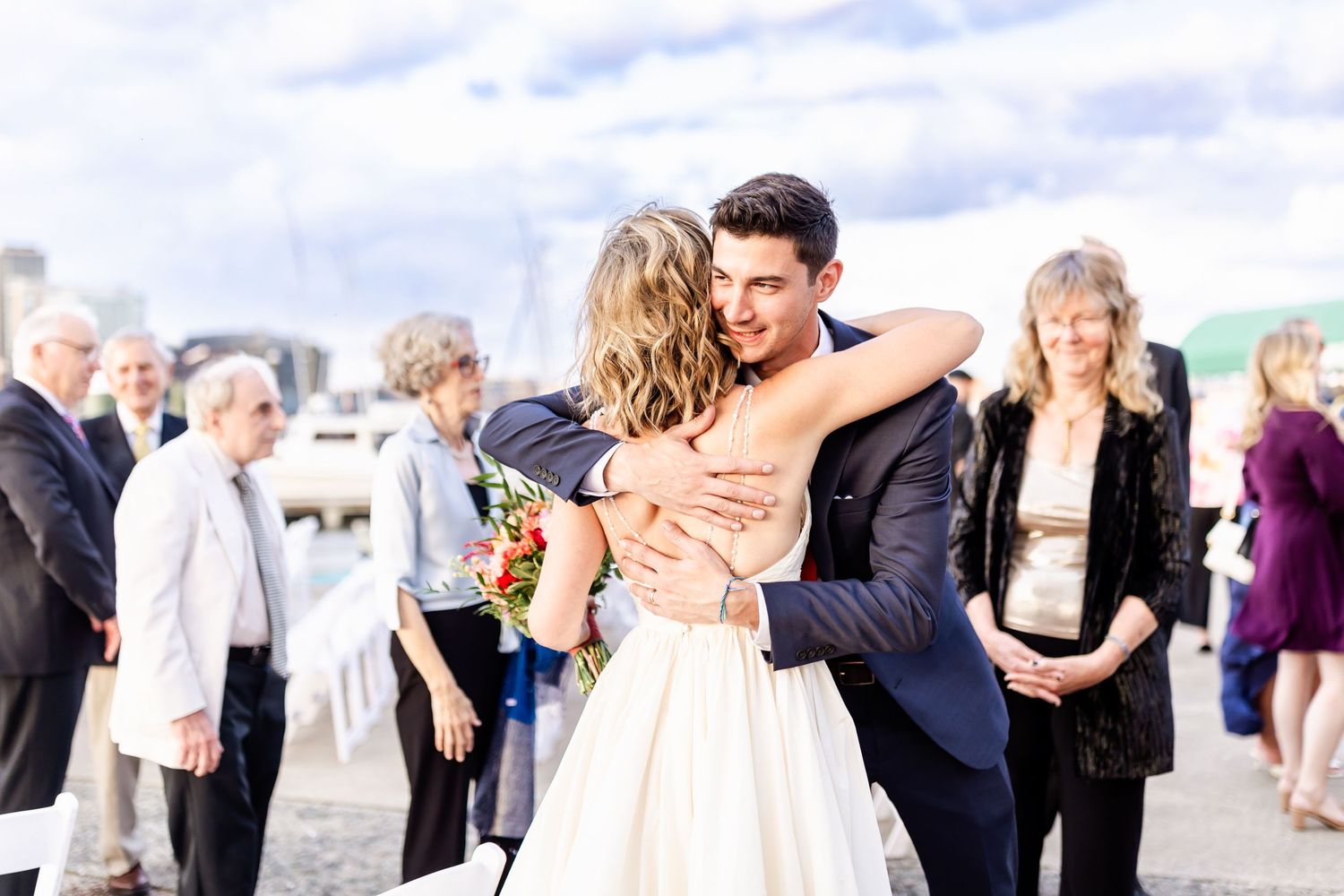 A tender embrace shared during an outdoor wedding celebration with guests looking on.