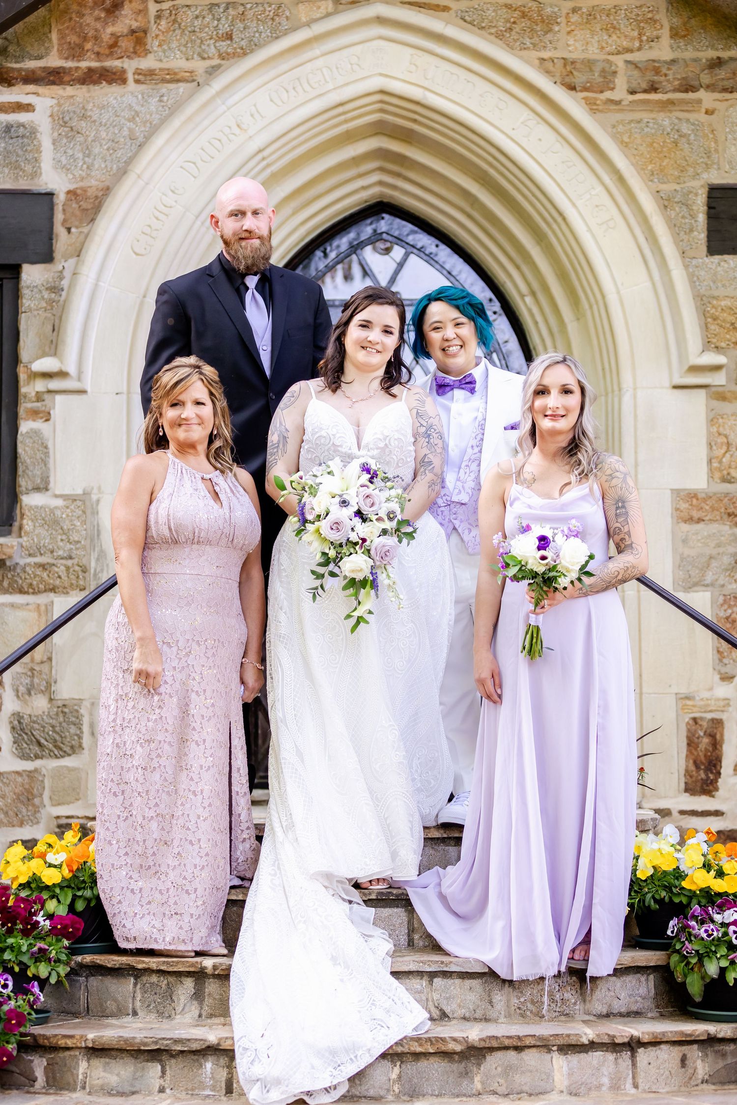 Formal wedding party portrait on church steps with gothic archway and purple floral arrangements.