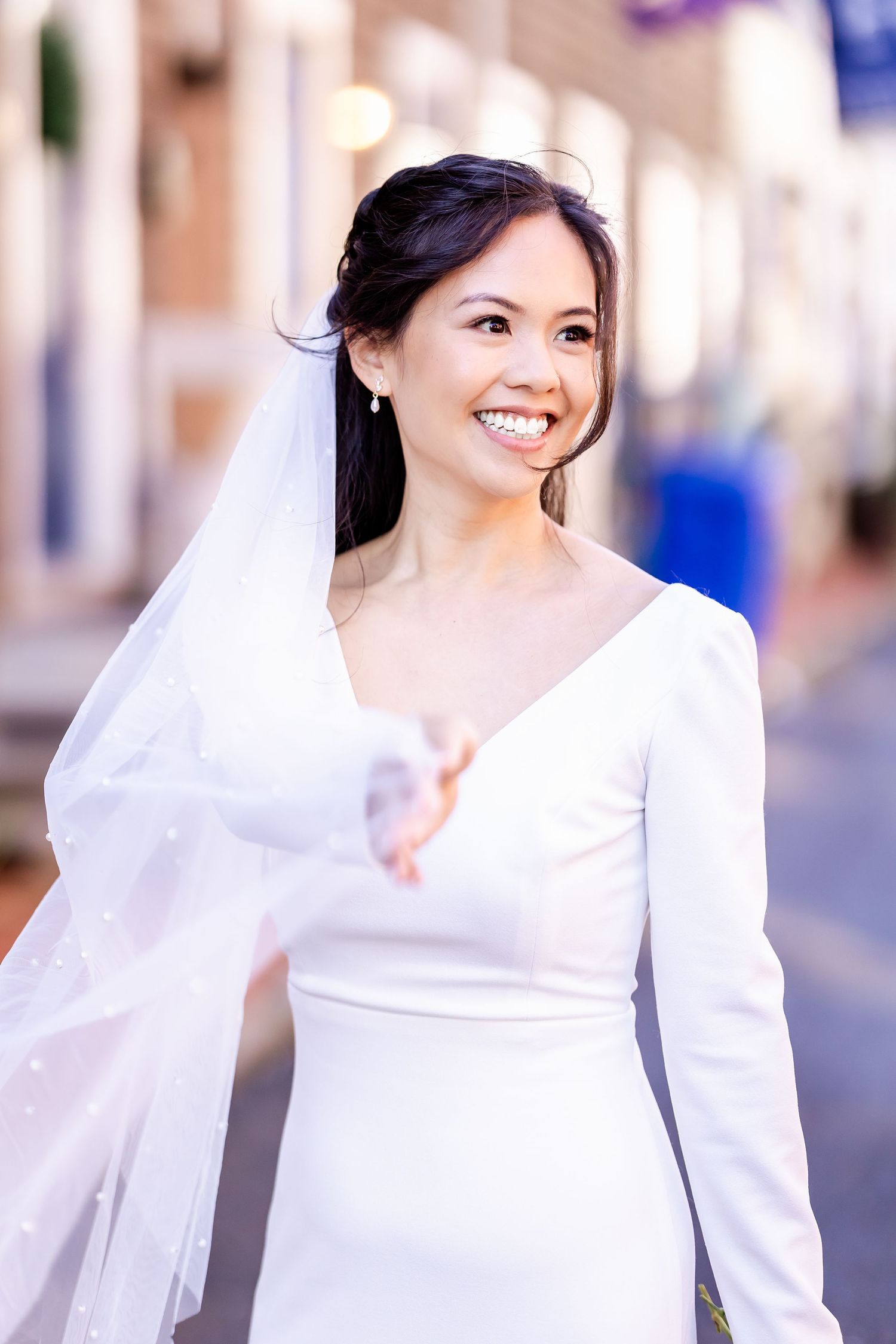 Radiant bride wearing elegant long-sleeve white dress and veil against urban background.