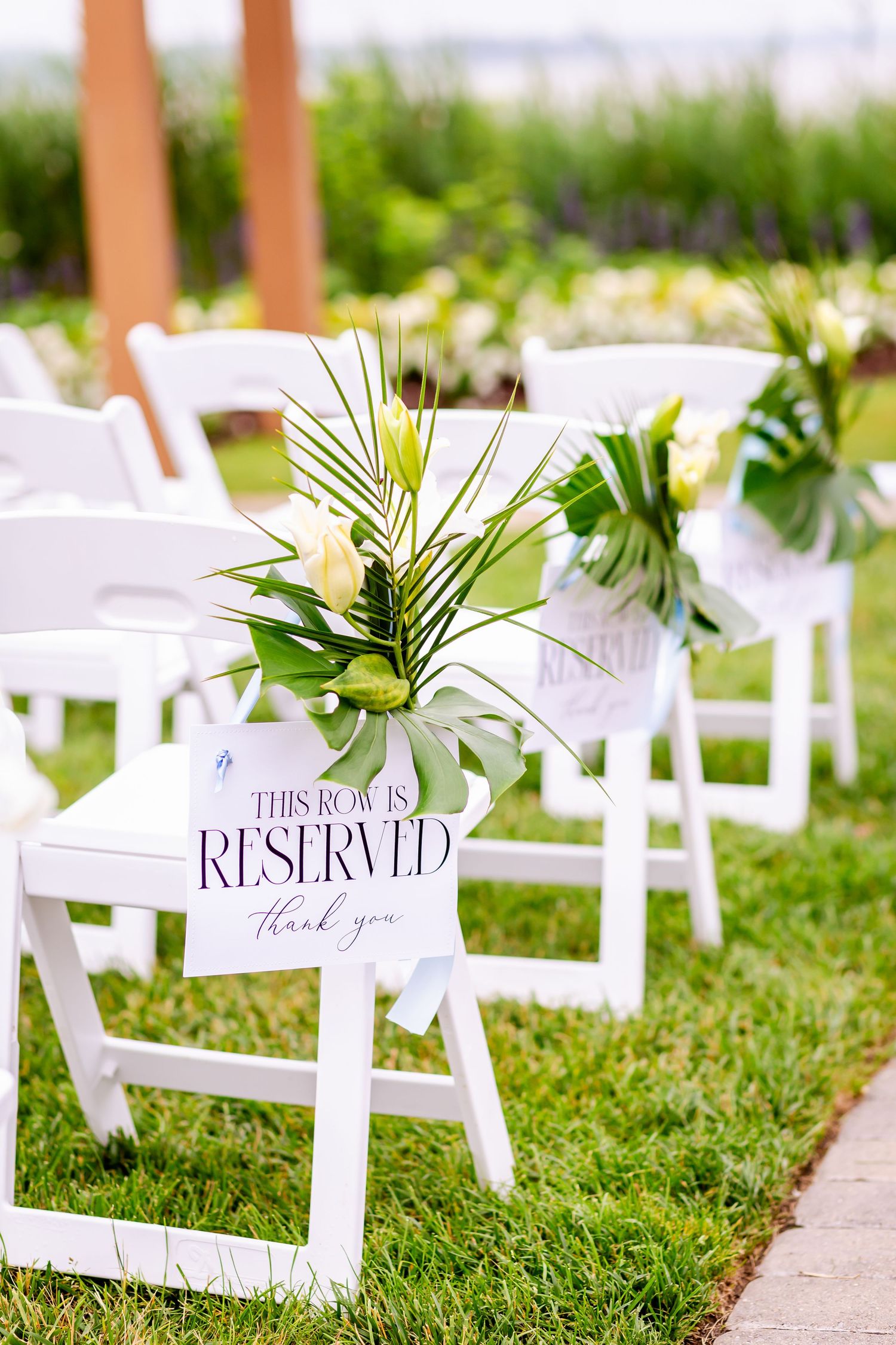 White folding chairs with reserved signs and tropical leaf decorations set up for an outdoor wedding ceremony.