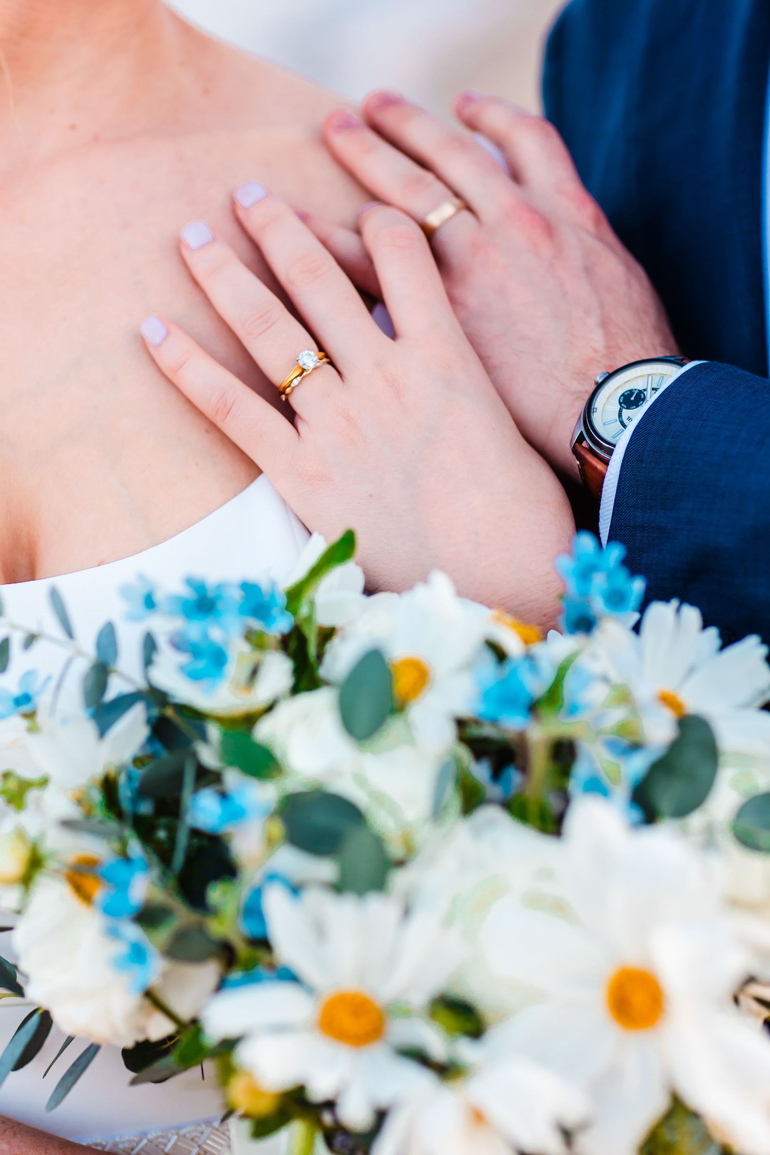 Wedding bouquet of white flowers and blue accents arranged with eucalyptus leaves and wedding rings visible.