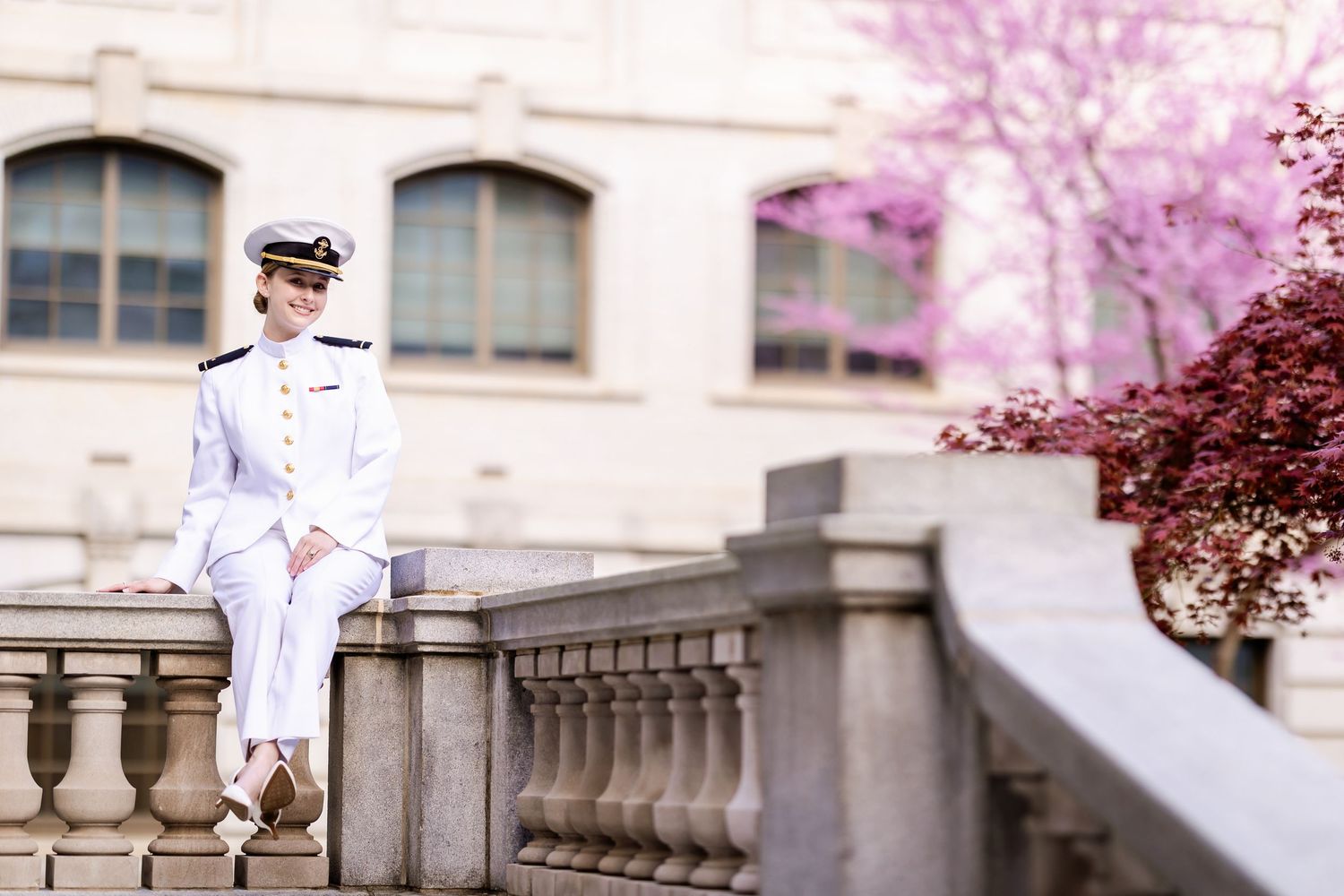 A person in a white naval uniform sits on a stone balustrade with pink cherry blossoms in the background.