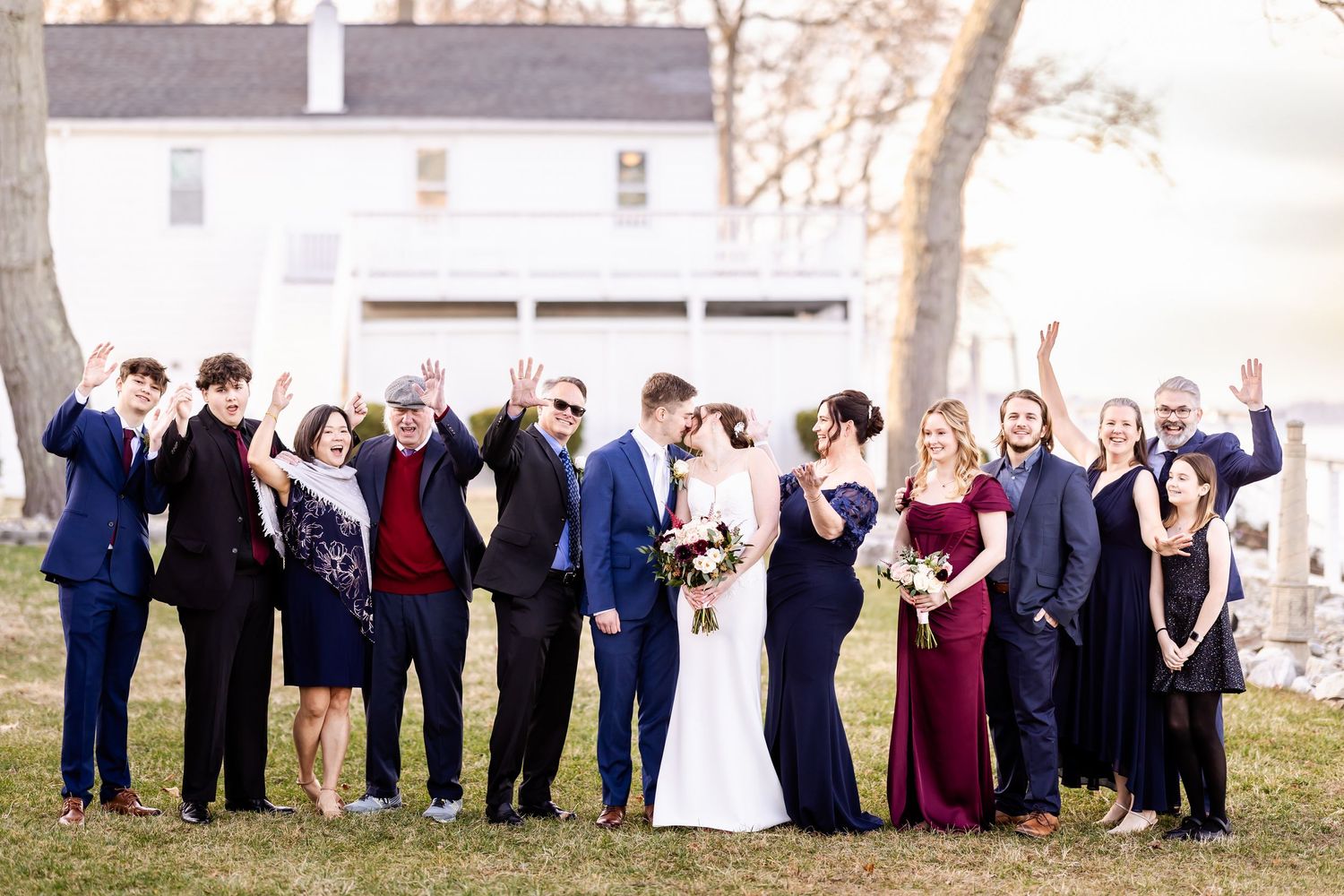 Large wedding group gathers on grassy lawn for formal outdoor portrait with historic building in background.
