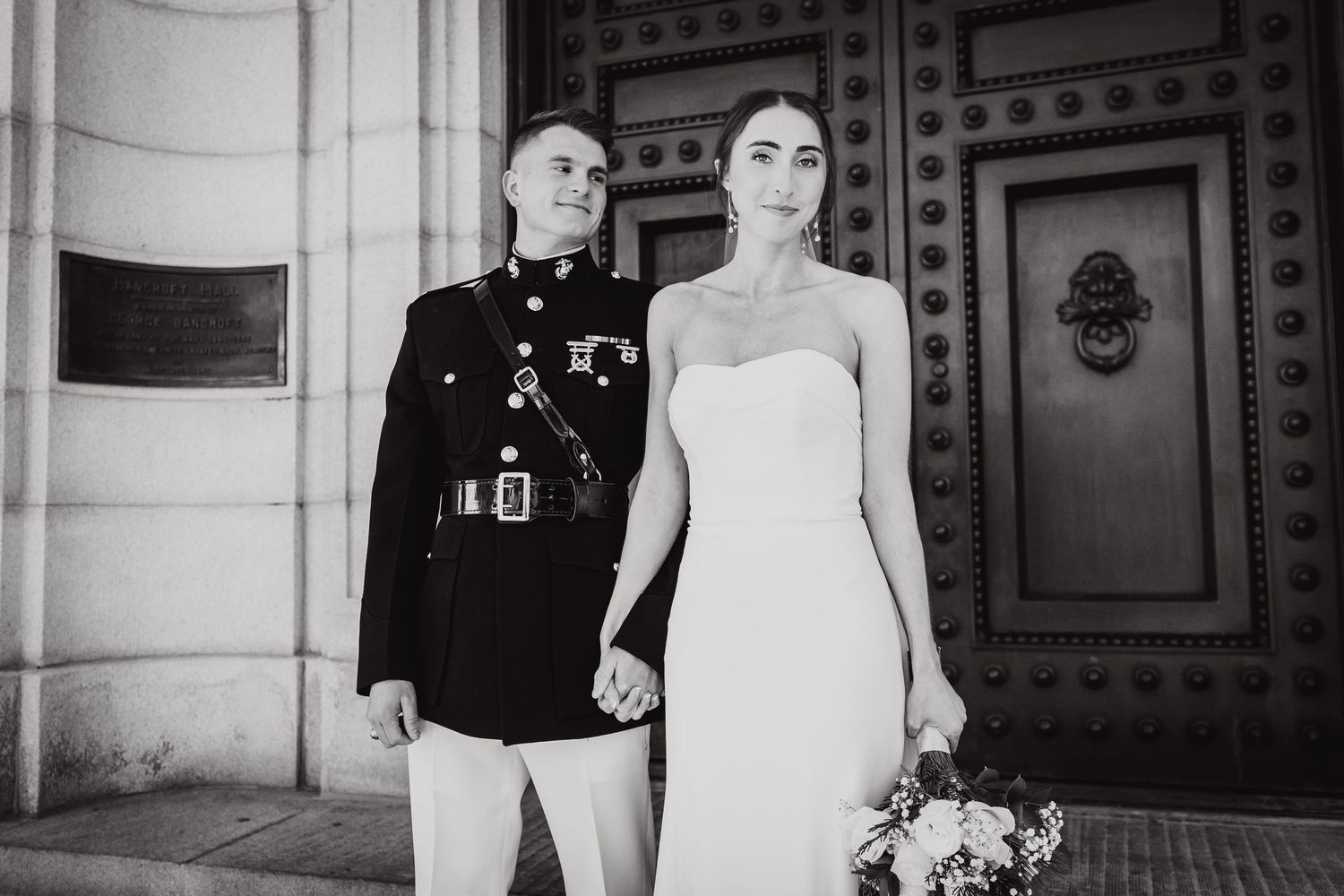 Elegant black and white wedding photo of military groom and bride in strapless dress holding hands outdoors.