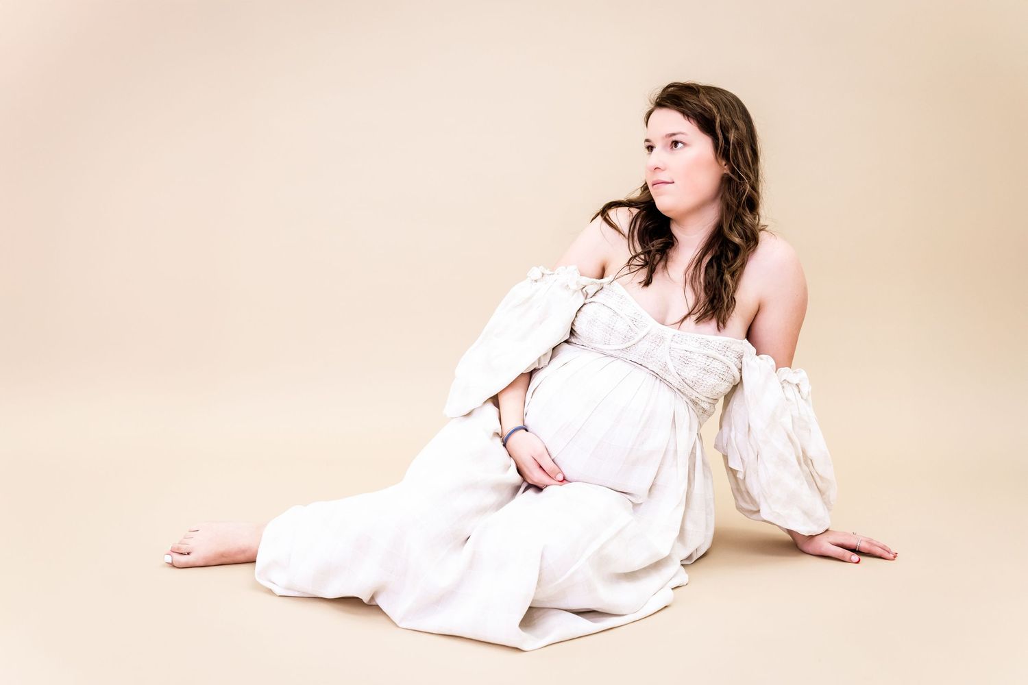 A maternity portrait featuring flowing white fabric and natural posing on a beige background.