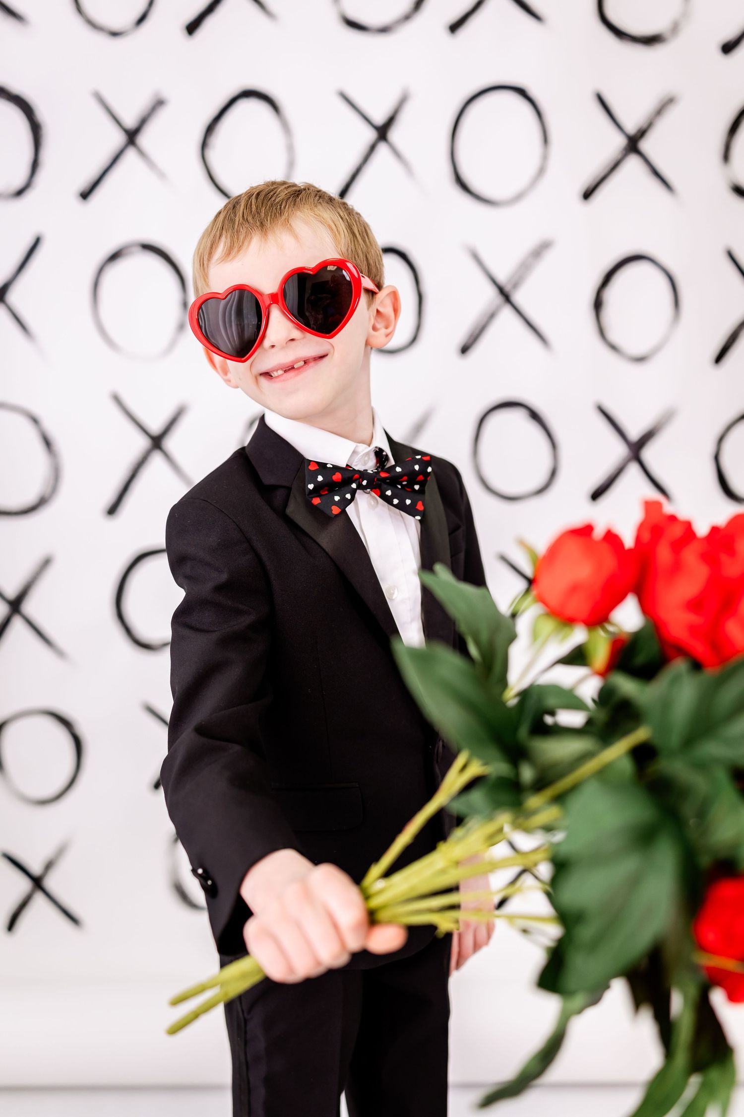 A young person in a black suit and heart-shaped red sunglasses holds red roses against an XO patterned background.