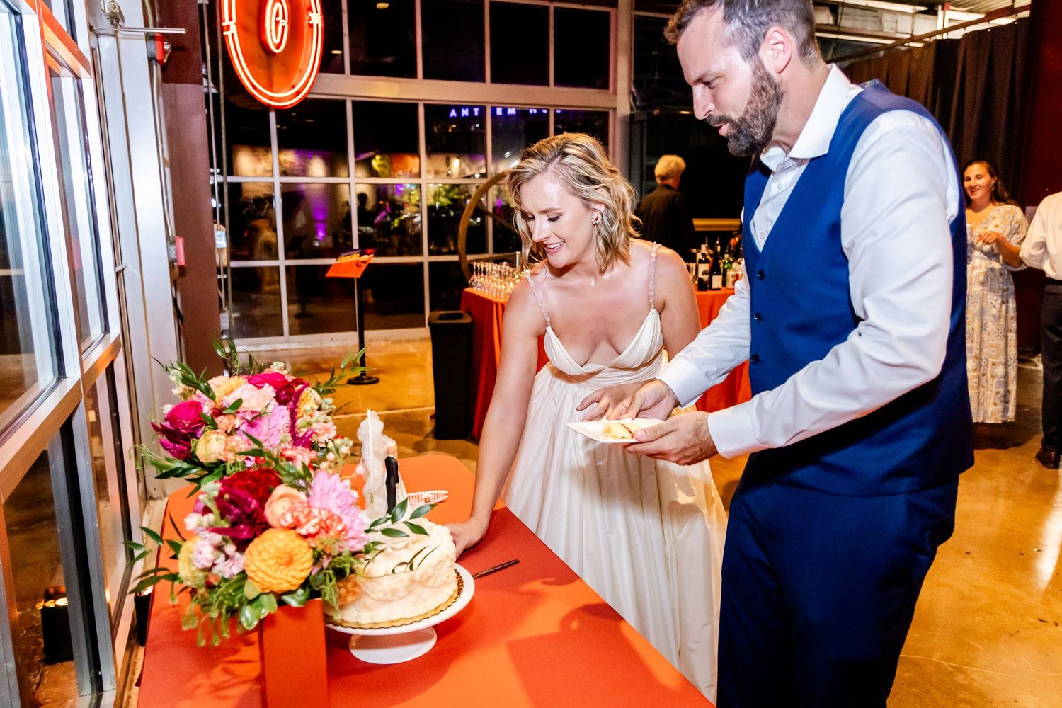 A bride and groom cut their wedding cake together at an indoor reception with pink and red floral decorations.