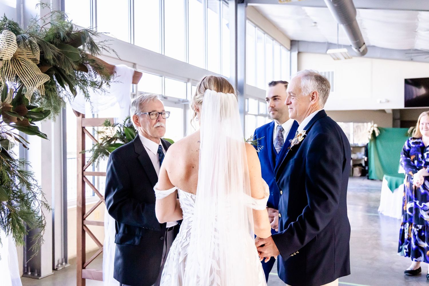 A wedding ceremony taking place in a bright indoor venue decorated with evergreen garlands.