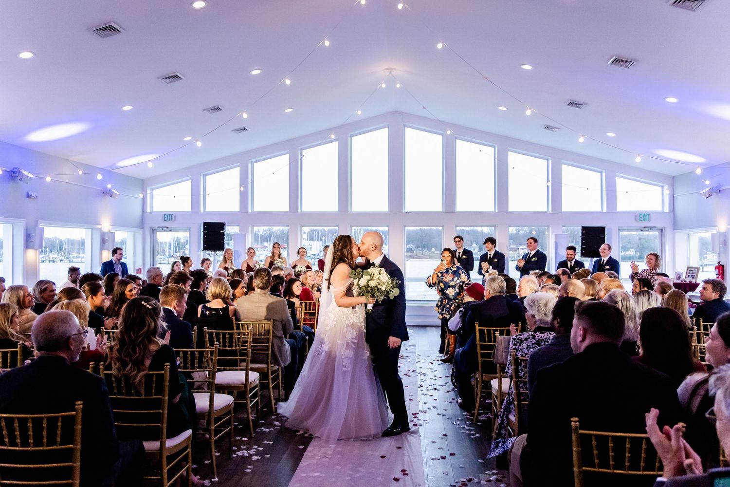 Wedding guests watch a ceremony in an elegant reception hall with white cathedral windows and pendant lights.