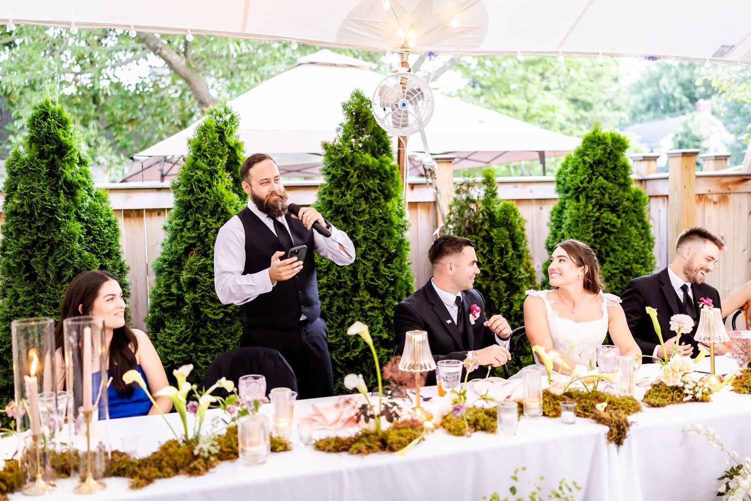 Wedding reception guests seated at decorated tables under a white tent with lush greenery.