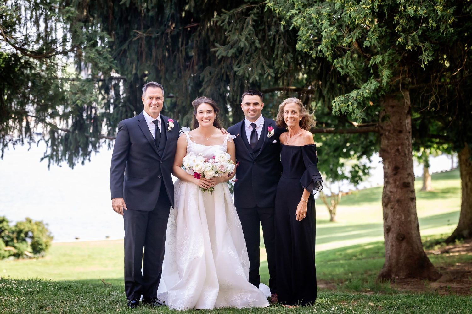 Wedding party members gather for formal photos on a grassy hill with evergreen trees.
