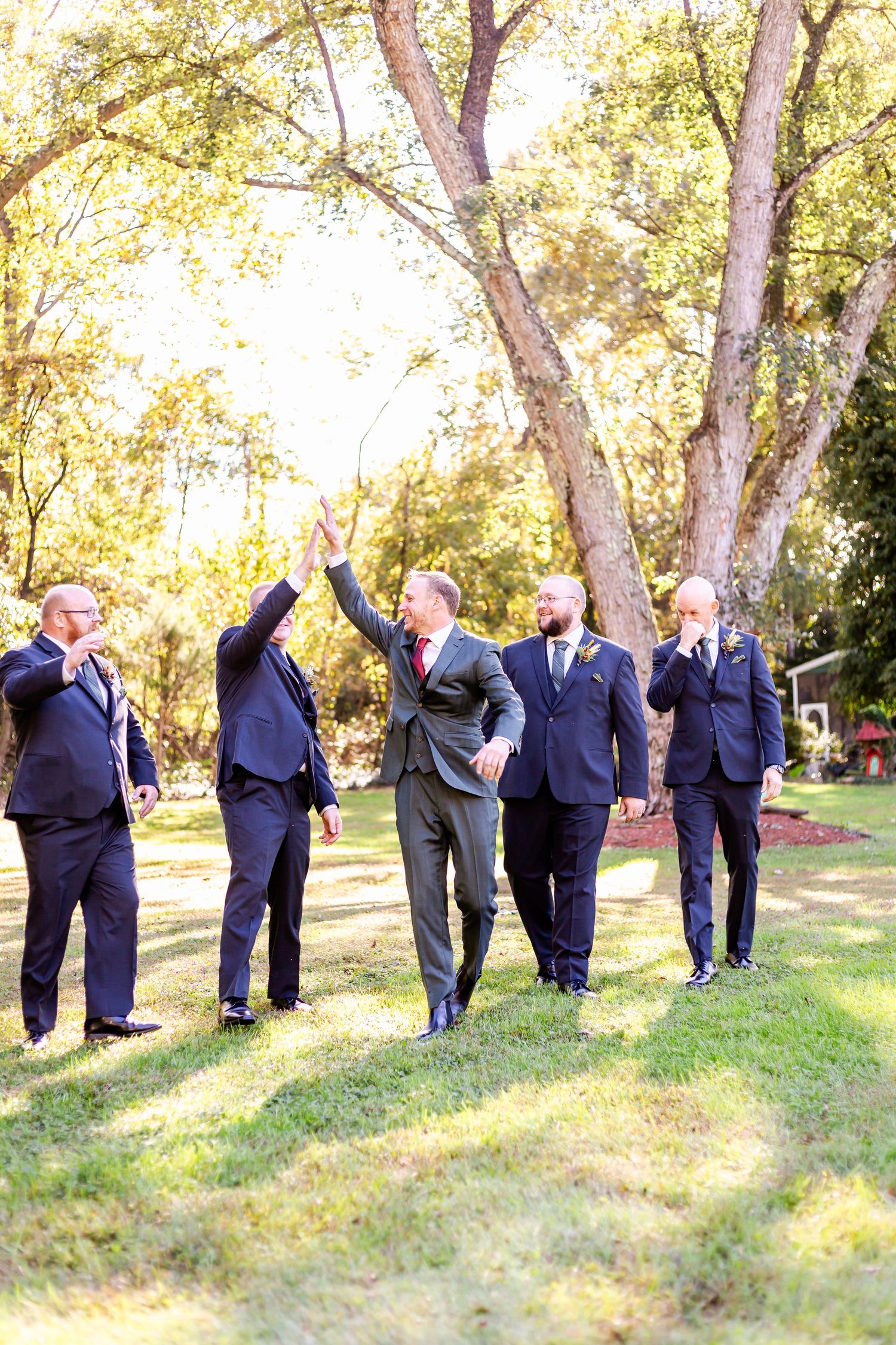 Groomsmen in navy suits pose playfully behind trees in a line at an outdoor wedding venue.