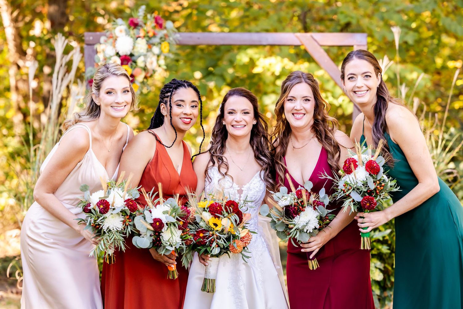 Wedding party in burgundy and emerald dresses holding colorful bouquets in outdoor autumn setting.