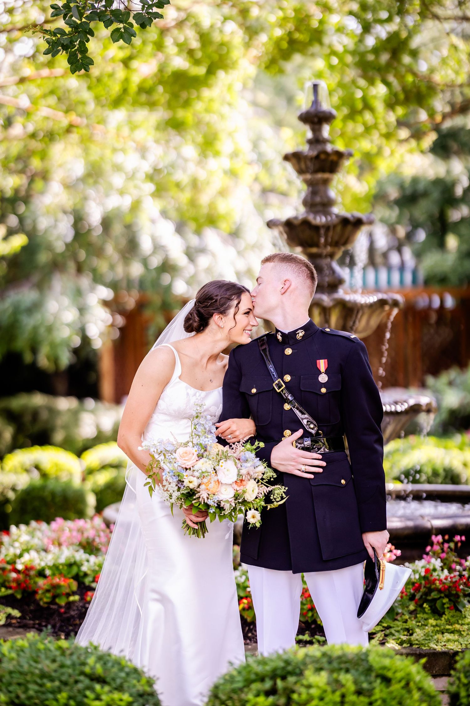 A romantic wedding couple shares intimate kisses in a garden setting with fountains and greenery.