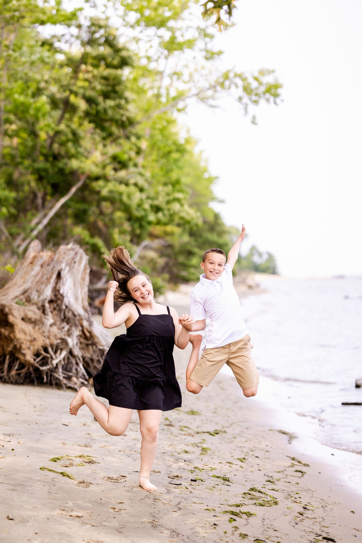 Two people jumping joyfully on a sandy beach with driftwood and trees in the background.