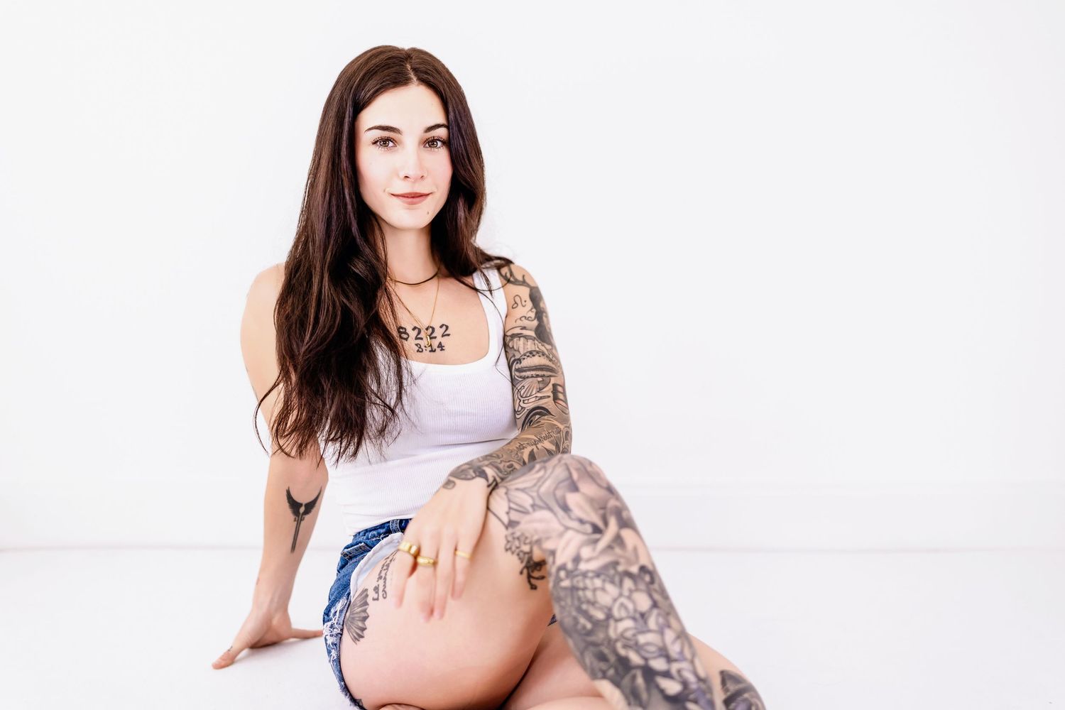 Person with long dark hair and tattoos wearing a tank top and shorts poses against white background.