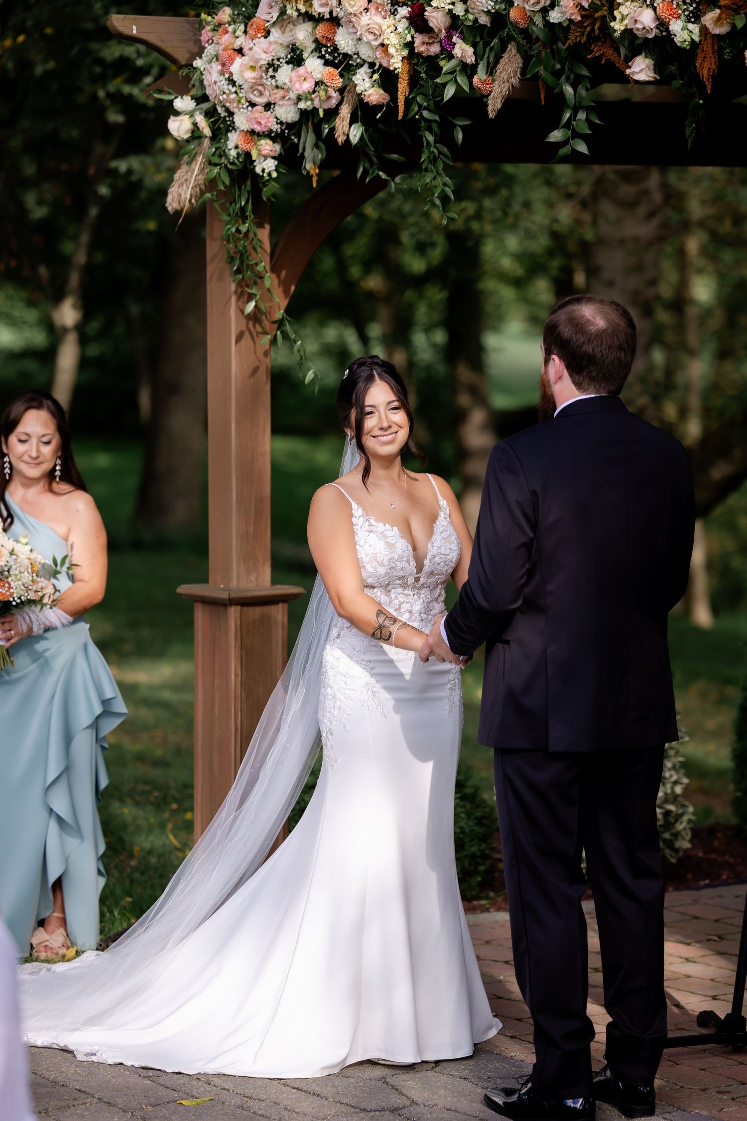 Bride in lace mermaid gown holds hands with groom at outdoor wedding ceremony beneath floral arch.