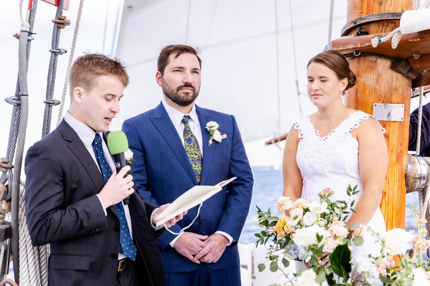 Officiant reads vows as groom in blue suit and bride in white lace dress stand together during outdoor wedding ceremony.