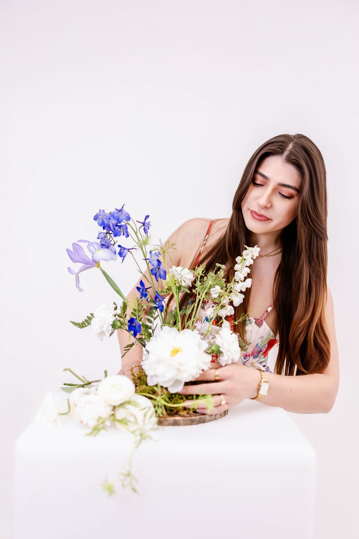 Young woman with long brown hair arranging a bouquet of blue irises and white peonies on a white pedestal.