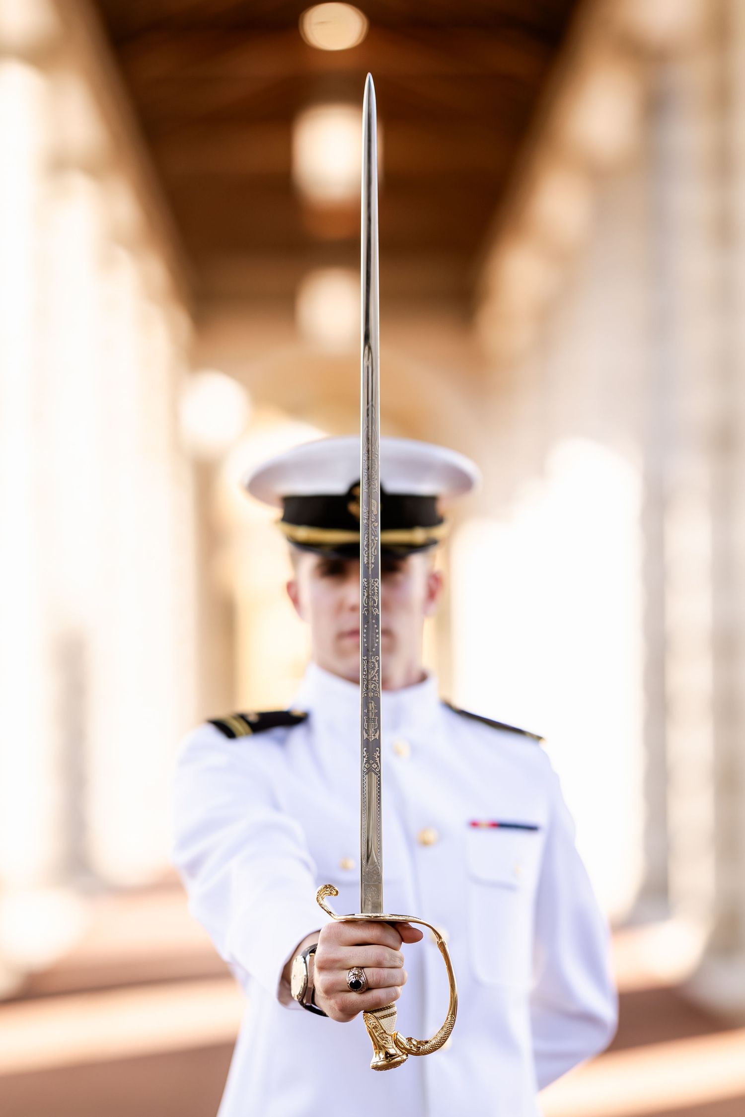 Naval officer in white dress uniform holding a ceremonial sword upright in a columned hallway.