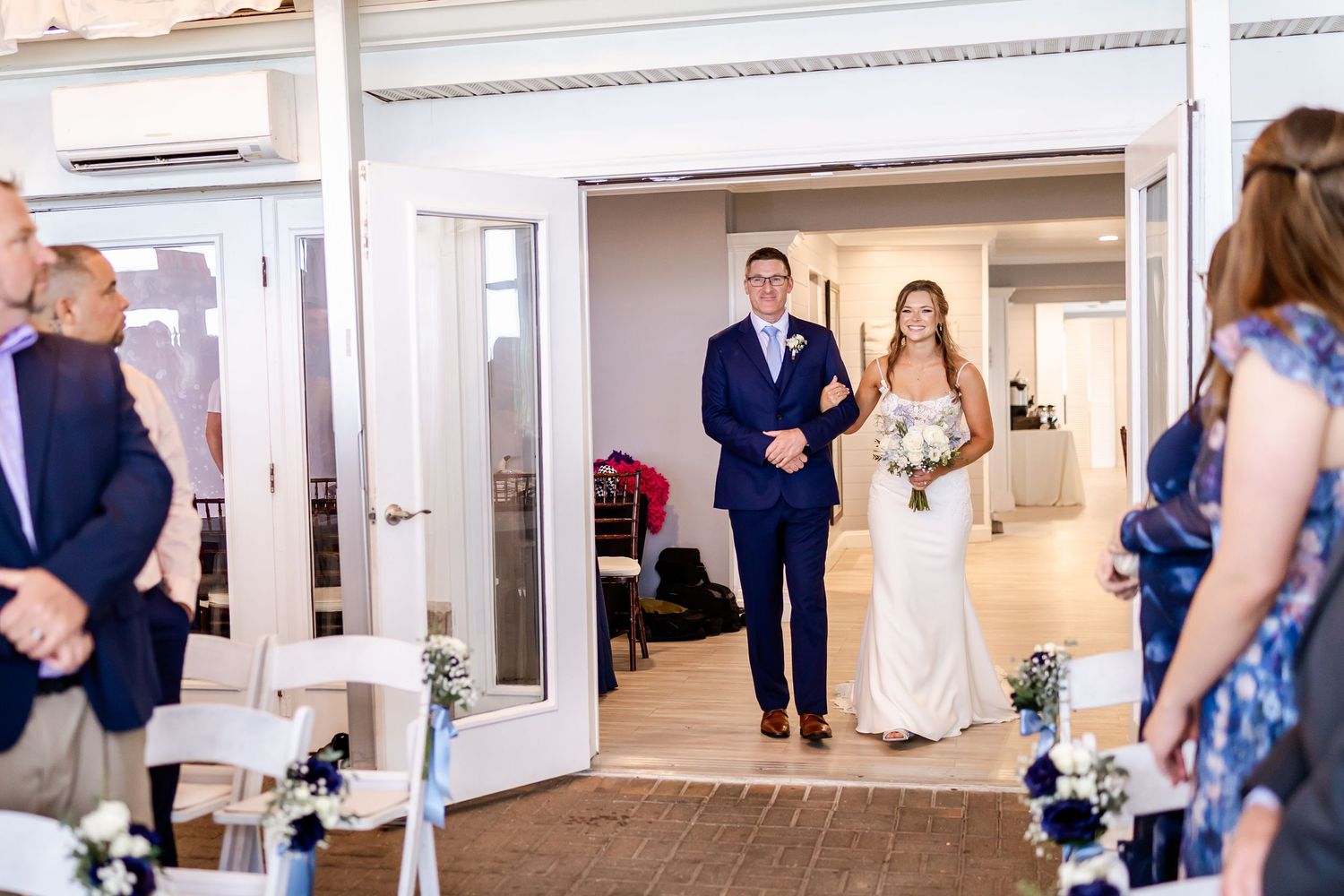 Wedding ceremony guests gather in an indoor venue with white decor.