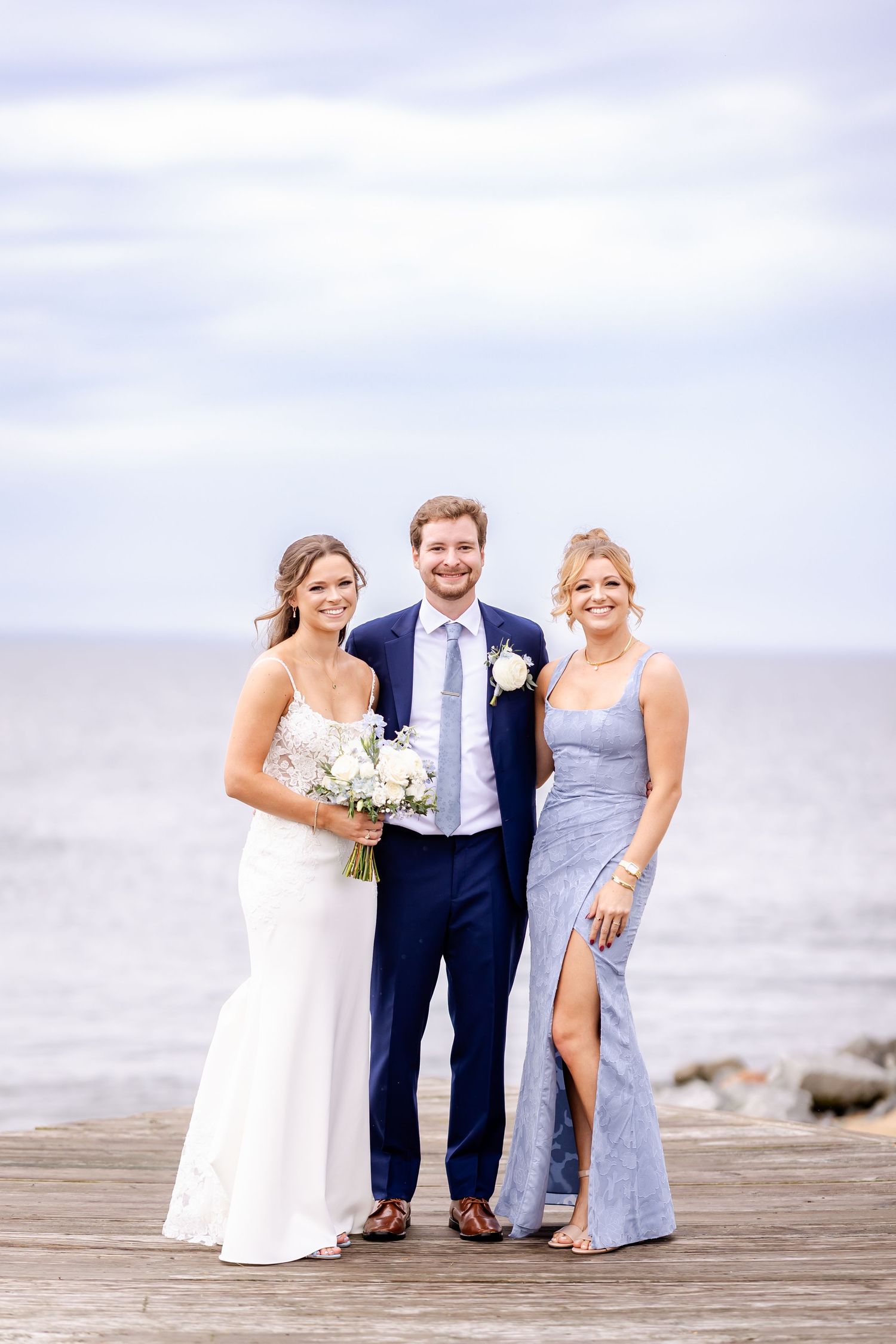 Wedding party poses playfully on a wooden boardwalk near the beach on an overcast day.