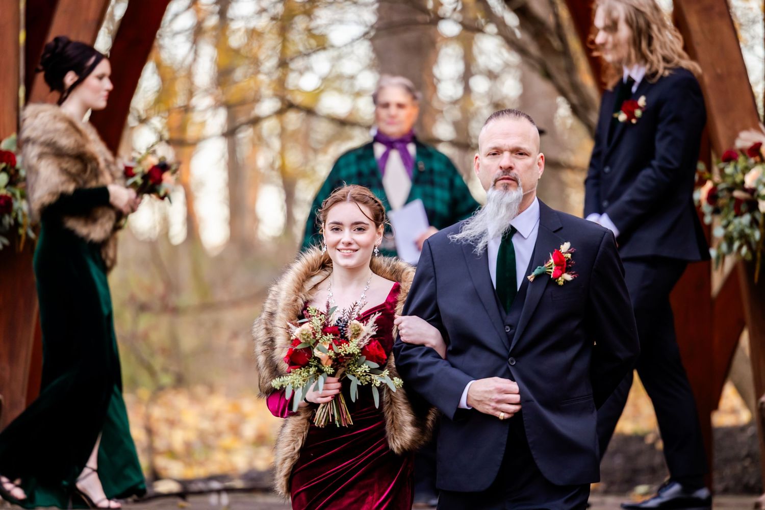 Wedding party members walk down an outdoor aisle with wooden arch and fall foliage backdrop.