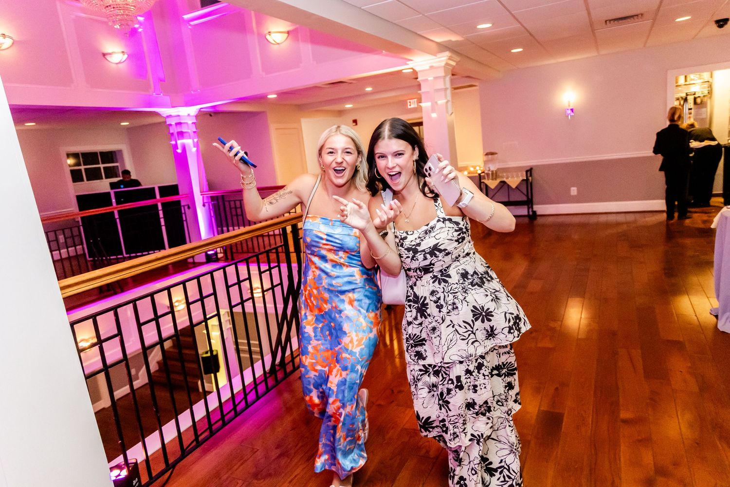People dancing and celebrating at an event in a banquet hall with pink lighting and hardwood floors.