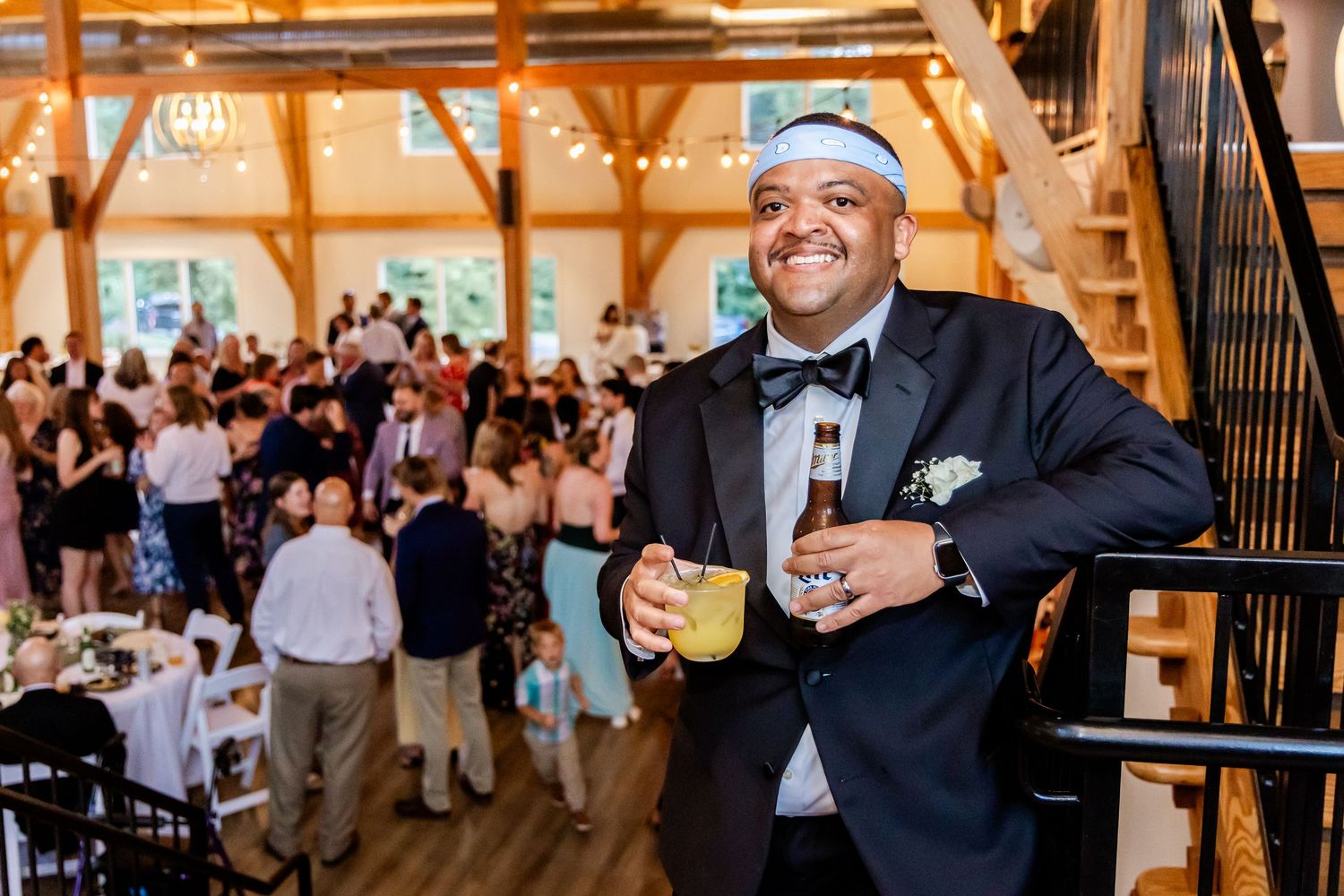 A festive wedding reception scene in a rustic wooden barn venue with warm lighting and guests mingling in the background.
