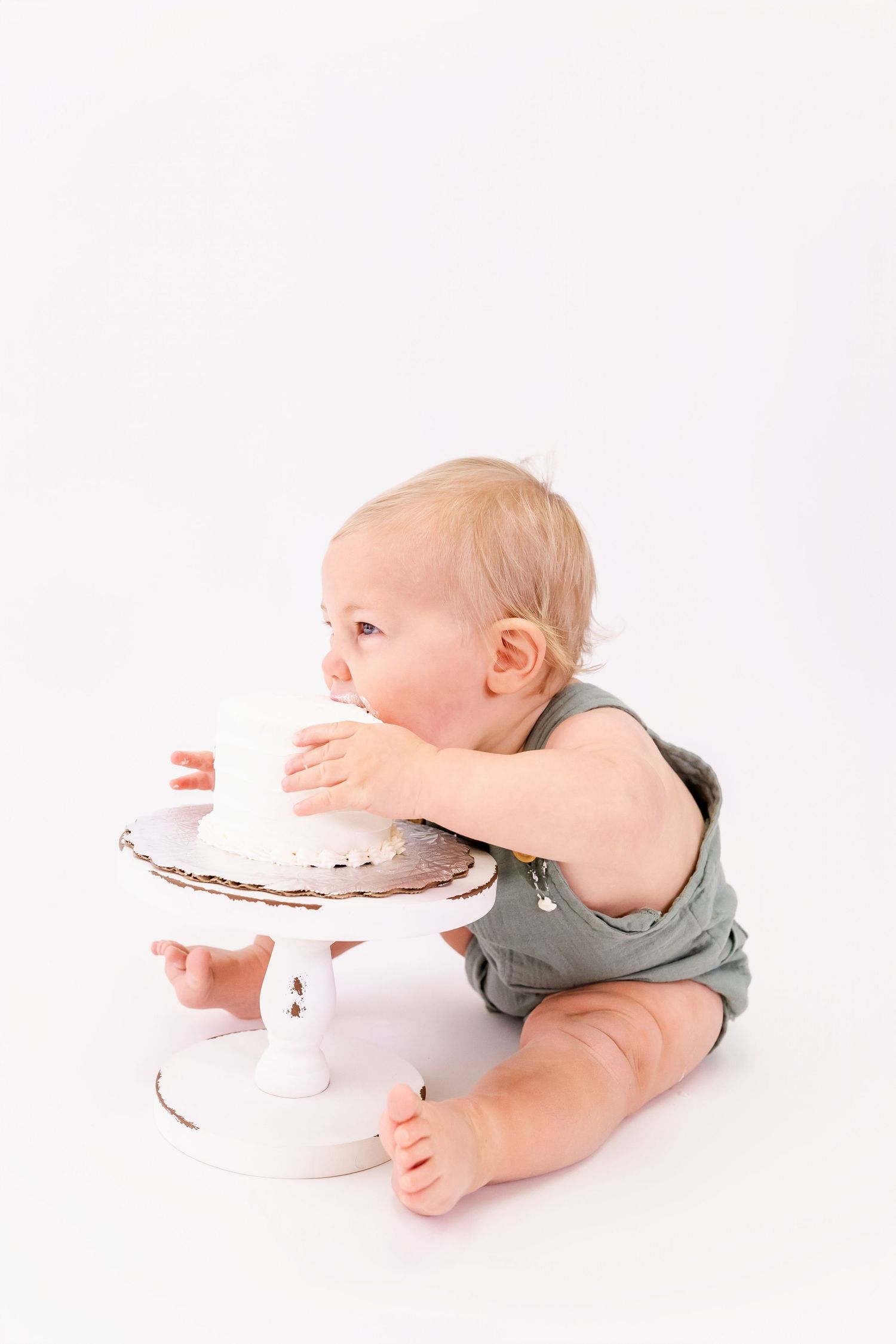 Baby in gray overalls reaching for white cake during photo session.