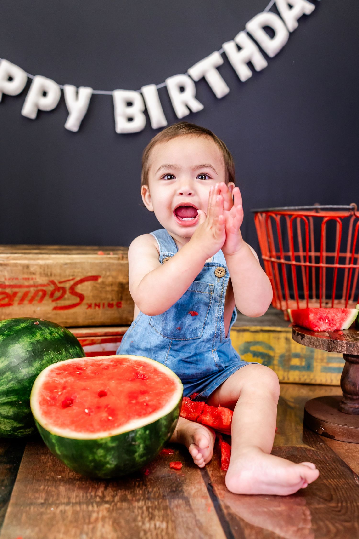 Excited toddler in denim overalls sitting next to large watermelon slice at birthday party setup.
